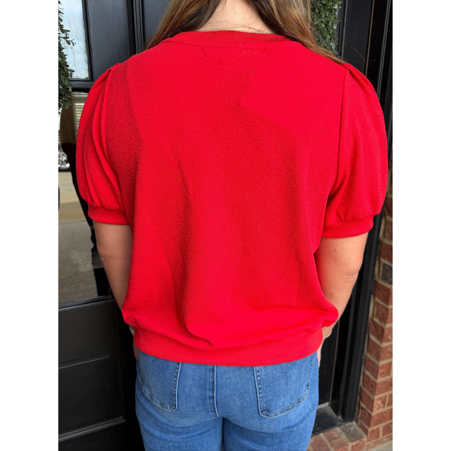 Person wearing a red blouse with puffed sleeves and blue jeans, standing in front of a door.