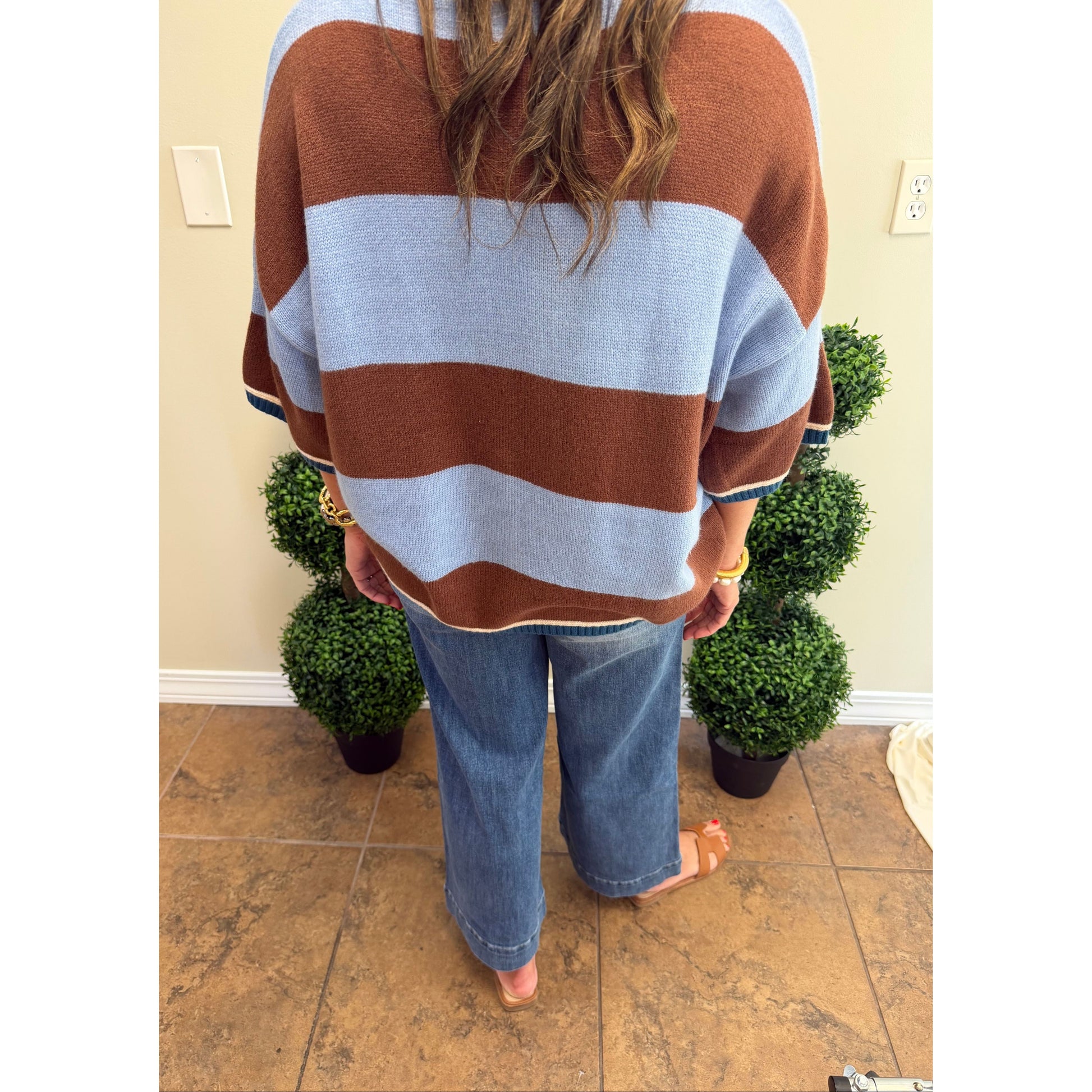 A person viewed from behind wearing a brown and light blue striped knit sweater, blue jeans, and tan sandals, standing indoors on a tiled floor with potted green plants against a beige wall.