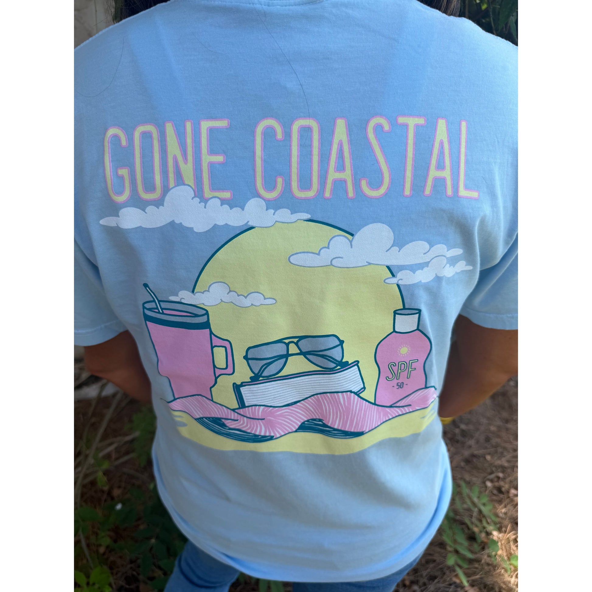 Light blue t-shirt with 'Gone Coastal' design featuring a sun, sunglasses, mug, book, and bottle on a person.