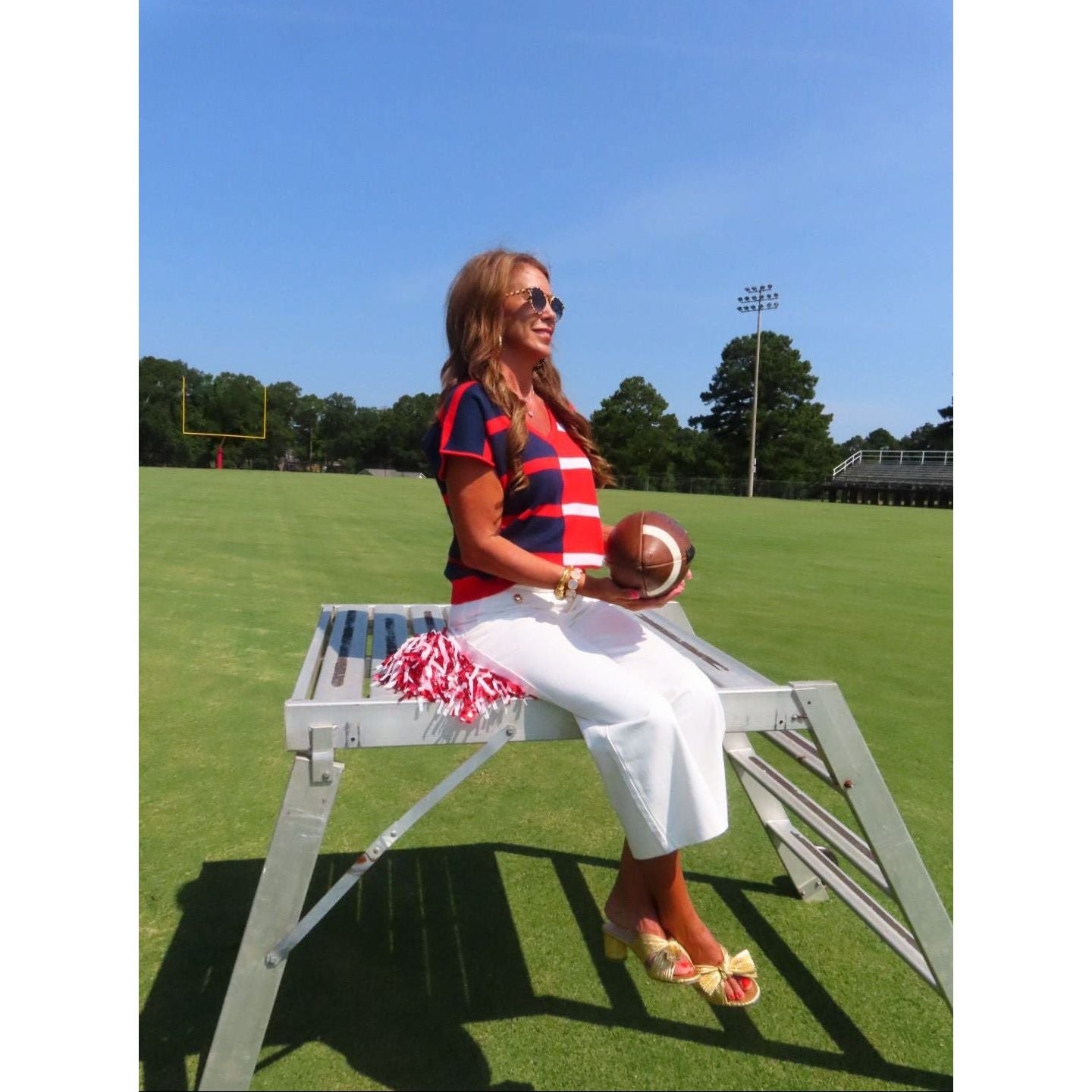 A woman sitting on a white metal bench on a grassy field, wearing a red, navy, and white checkered short-sleeve top, white pants, and holding a football.