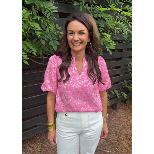Woman wearing a pink patterned blouse and white pants sitting on a bench with greenery around.
