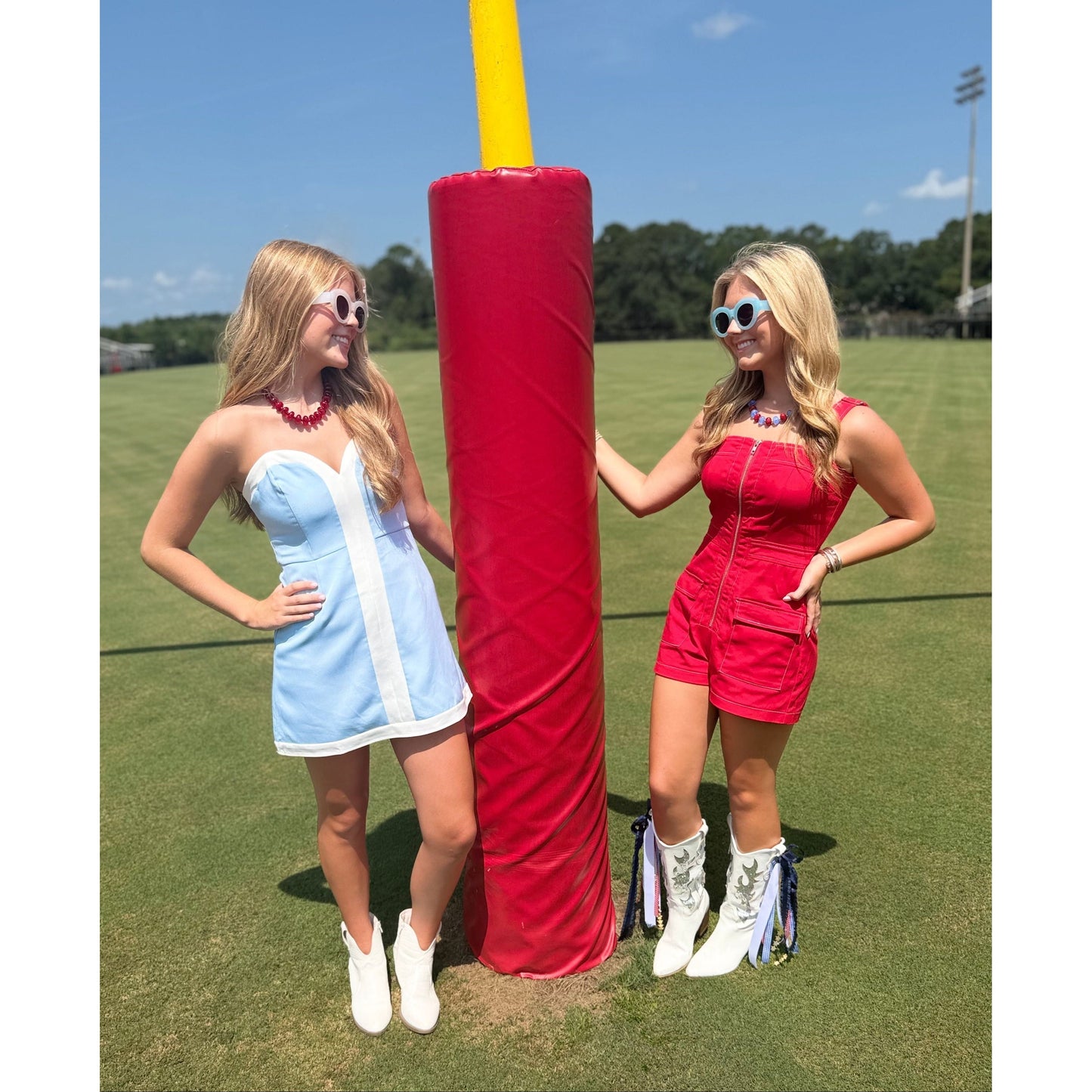 Two women posing outdoors on a grassy field in front of a large red and yellow football goal post. The woman on the left wears a light blue and white mini dress with white cowboy boots.