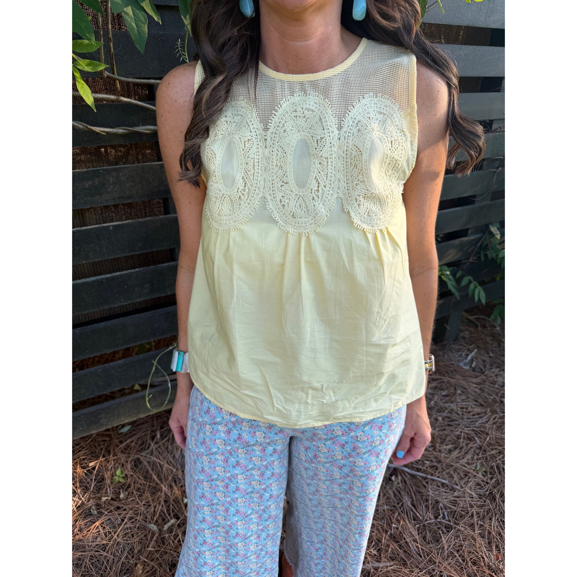 Woman wearing a light yellow sleeveless top with lace details and floral pants against a wooden fence background.
