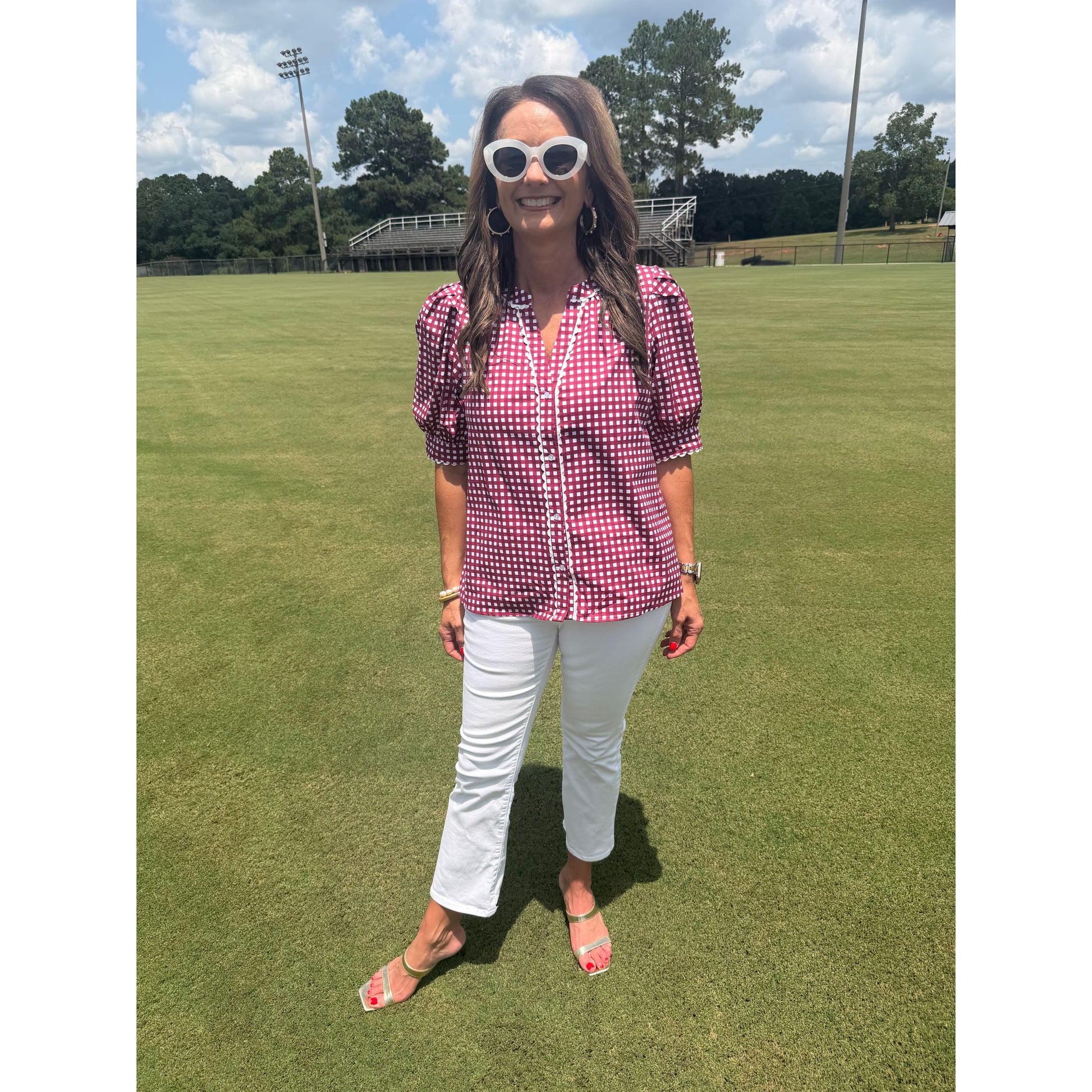 A woman stands on a green grass field wearing a pink and white gingham short-sleeve button-up shirt, white pants, and white sunglasses.