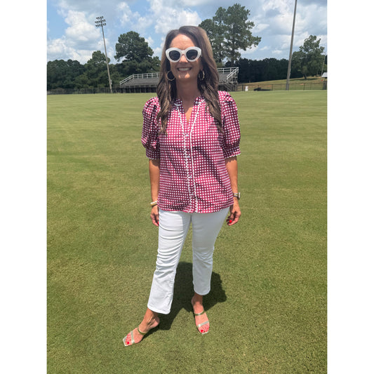 A woman stands on a green grass field wearing a pink and white gingham short-sleeve button-up shirt, white pants, and white sunglasses.