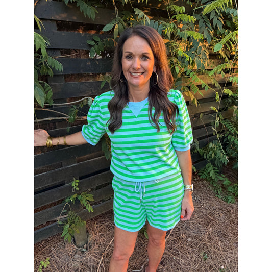 Woman wearing a green and white striped outfit standing against a wooden fence with plants.