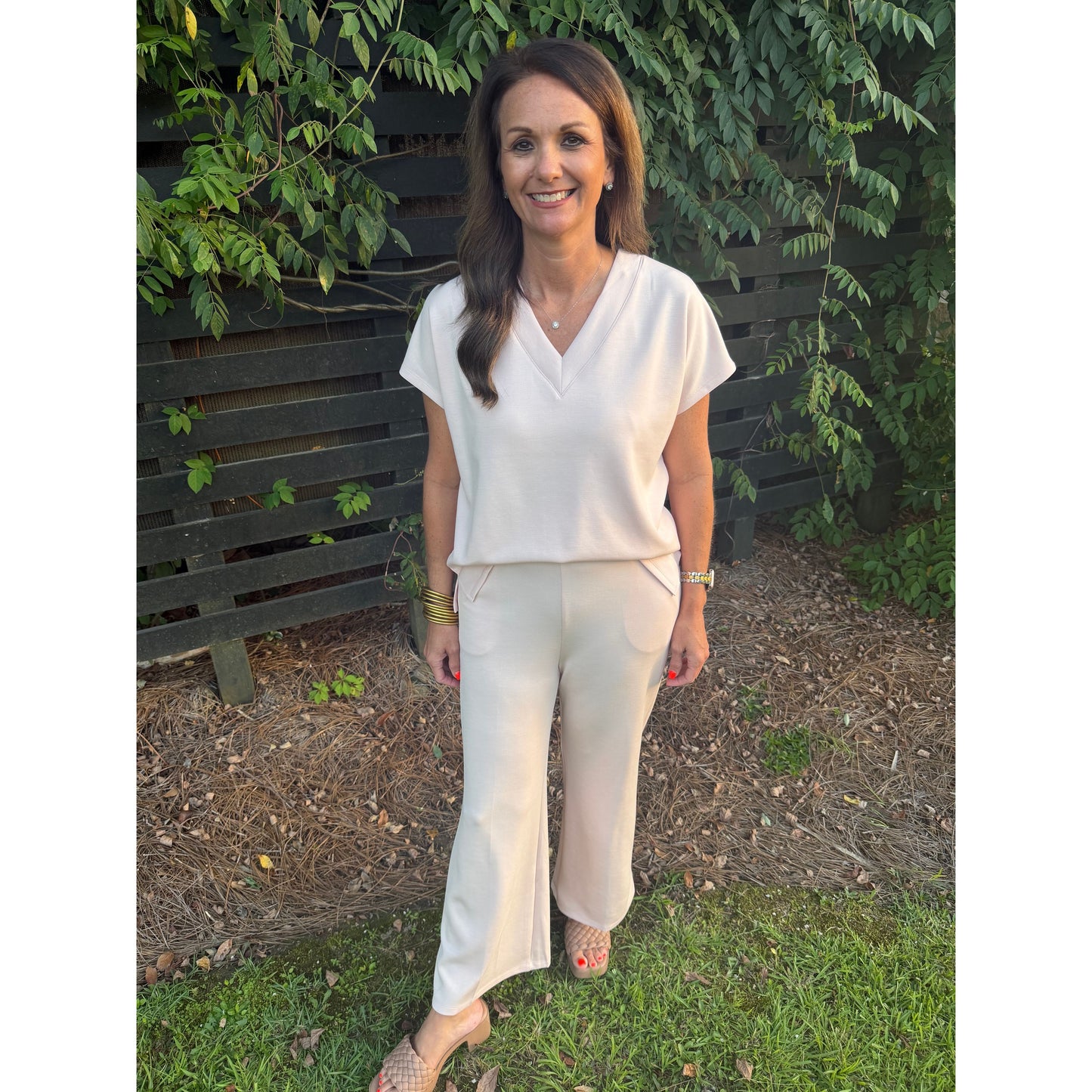 A woman wearing a cream-colored short-sleeve V-neck top and matching wide-leg trousers, standing outdoors on grass in front of a dark wooden fence and green foliage.