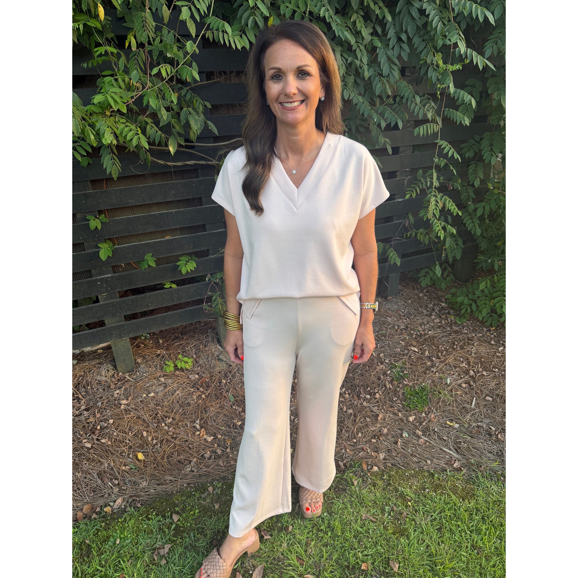 A woman wearing a cream-colored short-sleeve V-neck top and matching wide-leg trousers, standing outdoors on grass in front of a dark wooden fence and green foliage.