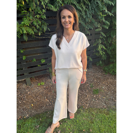 A woman wearing a cream-colored short-sleeve V-neck top and matching wide-leg trousers, standing outdoors on grass in front of a dark wooden fence and green foliage.