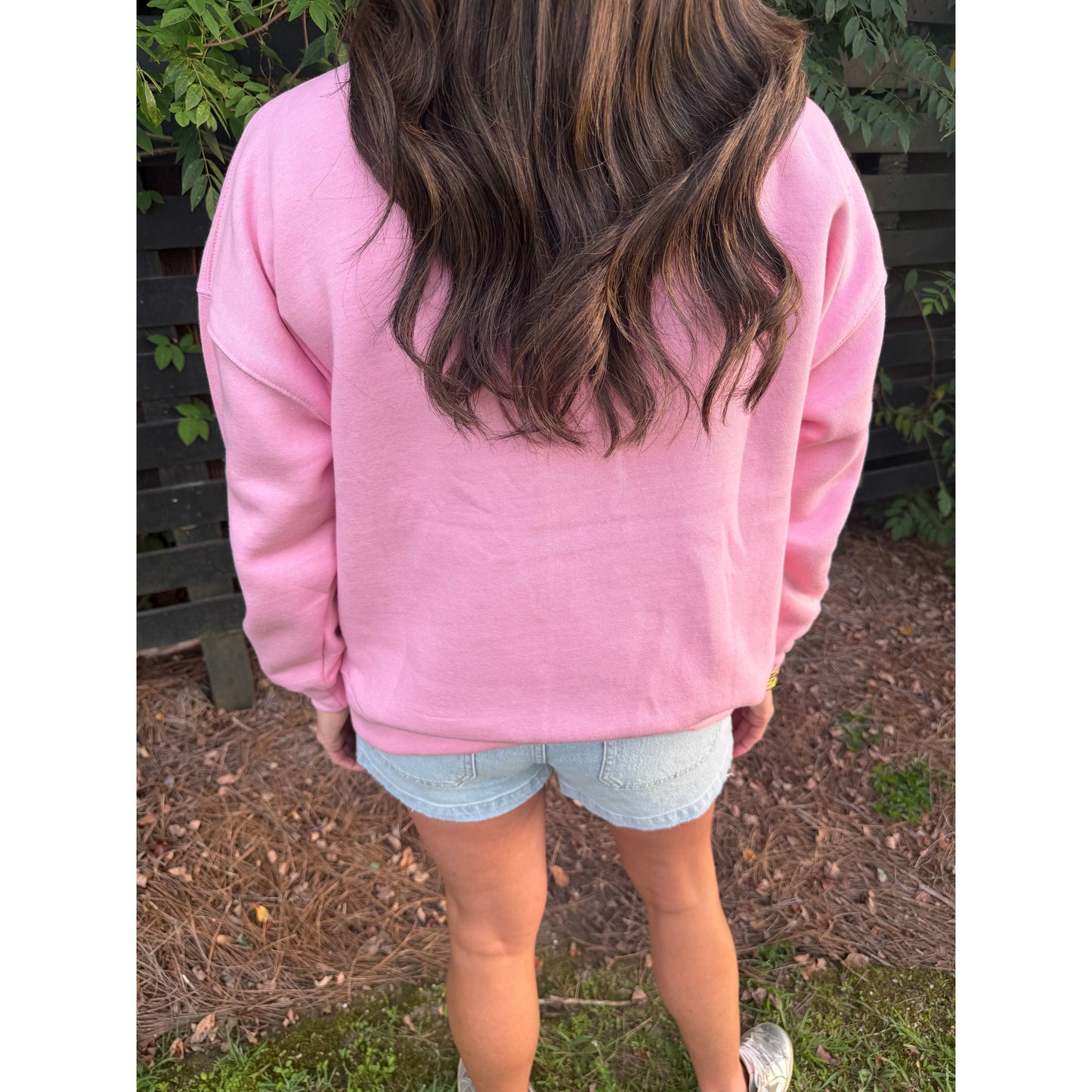 A person viewed from behind wearing a pink crewneck sweatshirt and light blue denim shorts, standing outdoors on a mulch and grass surface with a dark fence and greenery in the background.