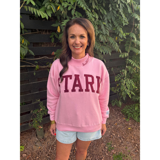 A woman wearing a pink crewneck sweatshirt with 'STARK' printed in maroon block letters across the chest, paired with light denim shorts, standing outdoors in front of a dark wooden fence and green foliage.