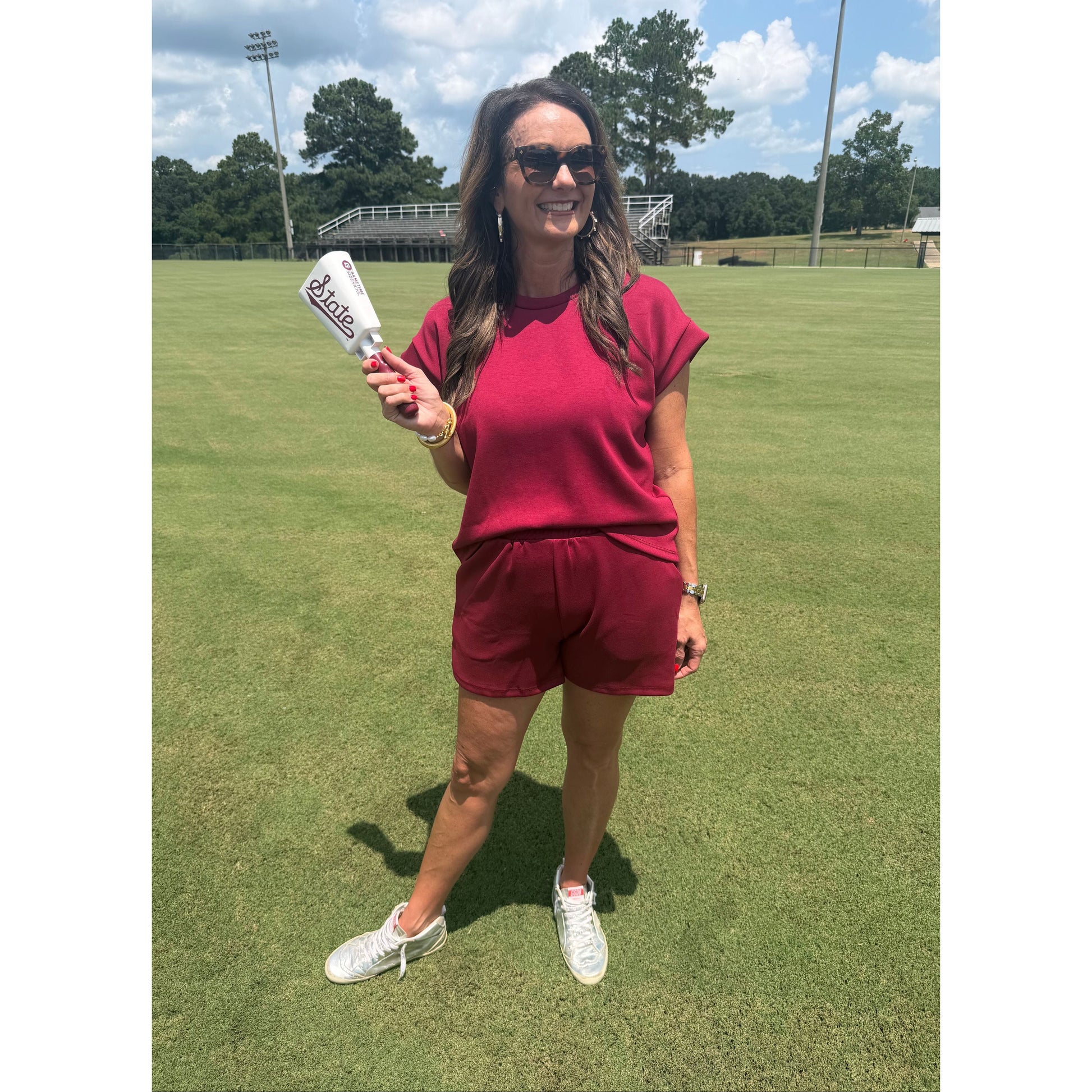A woman wearing a matching burgundy short-sleeve top and shorts set stands on a green grass field, holding a white bottle with a red label. She is wearing white sneakers and sunglasses.