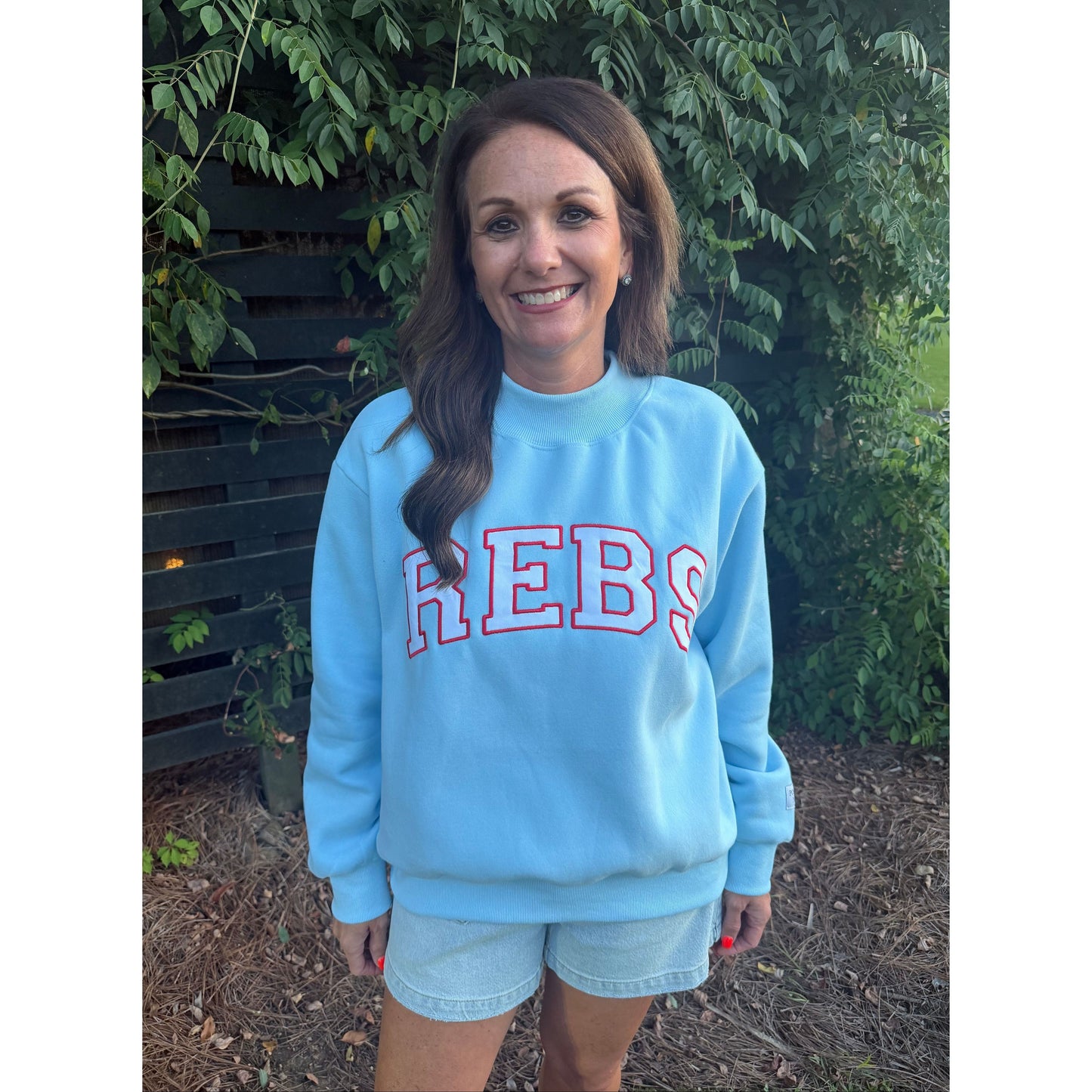 A woman wearing a light blue crewneck sweatshirt with 'REBS' printed in red varsity letters, standing outdoors in front of green foliage and a wooden bench.