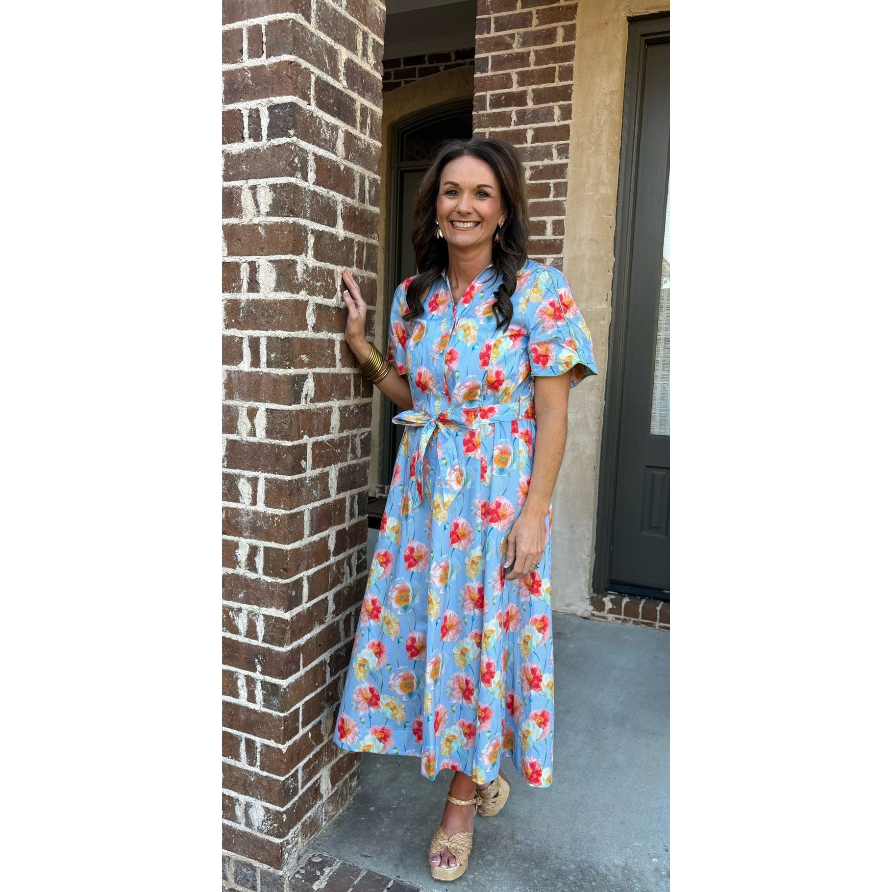 Woman in a floral dress standing on a porch next to a brick wall.