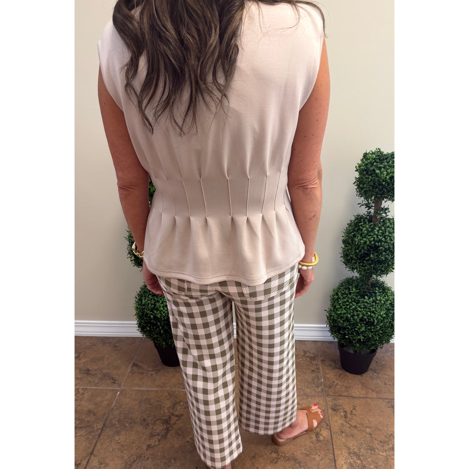 A woman viewed from behind wearing a sleeveless light beige peplum top and brown-and-beige gingham wide-leg pants, standing on a tiled floor with potted plants in the background.