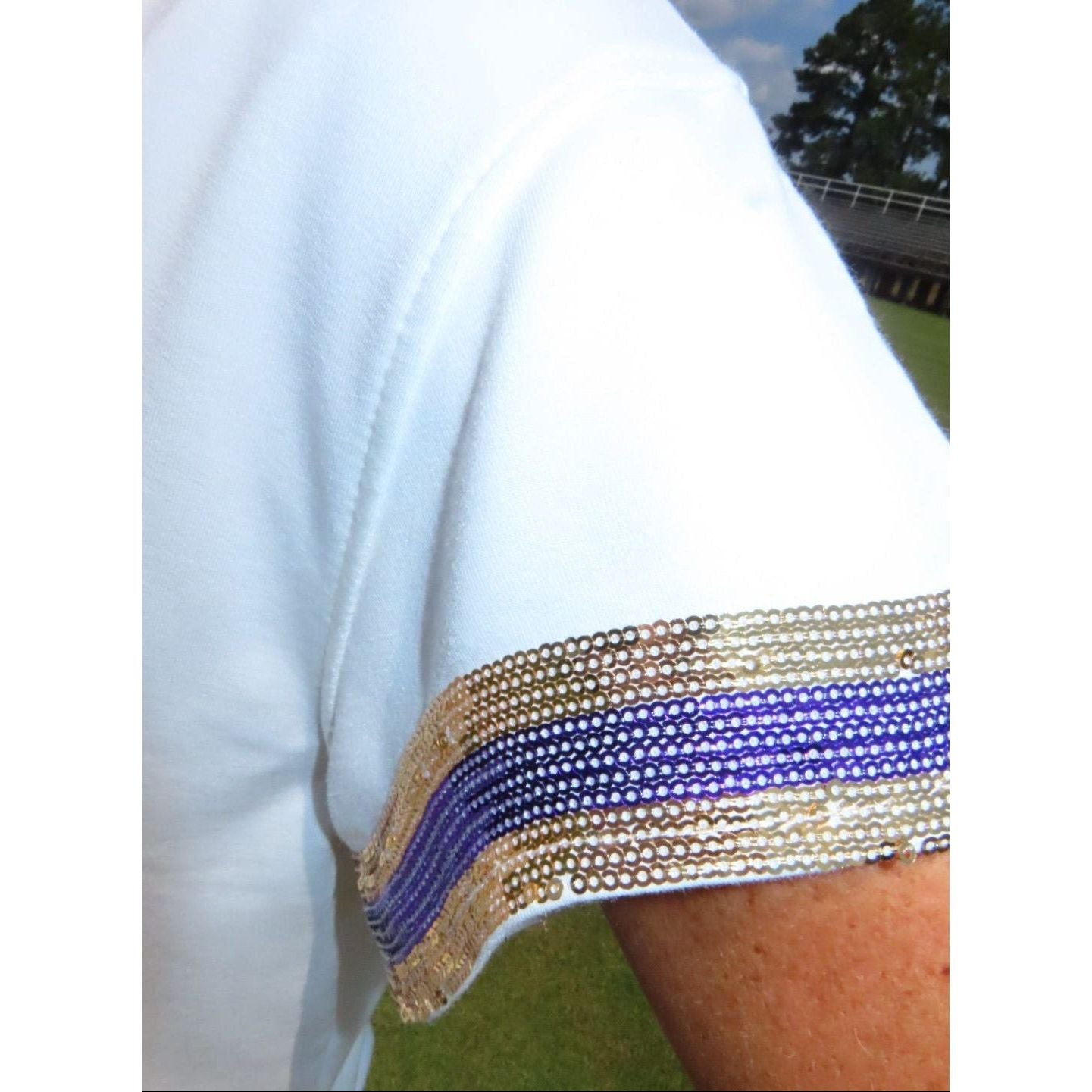 Close-up of a white short-sleeve garment with a decorative beaded sleeve cuff featuring horizontal stripes of gold, purple, and silver beads, worn on a person's arm outdoors.
