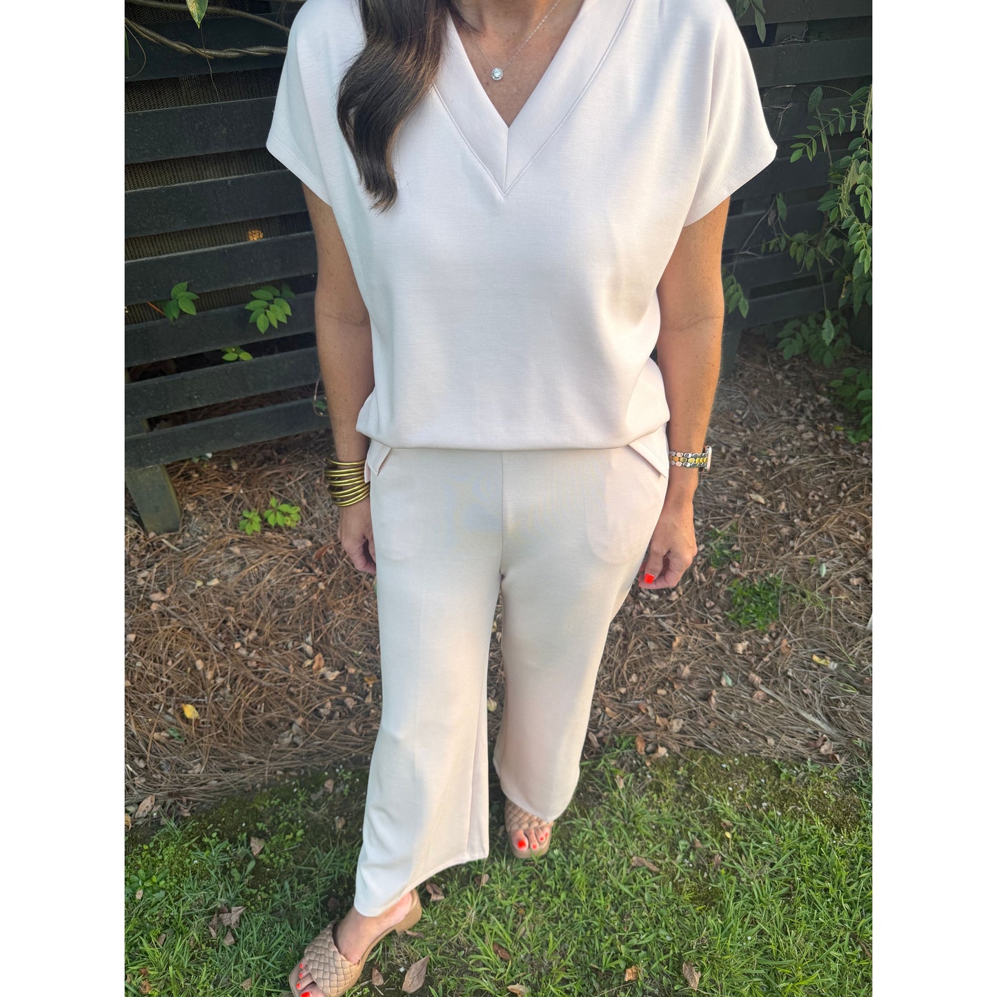 A woman wearing a white short-sleeve V-neck top and matching white wide-leg pants, photographed outdoors on grass and mulch next to a bench.