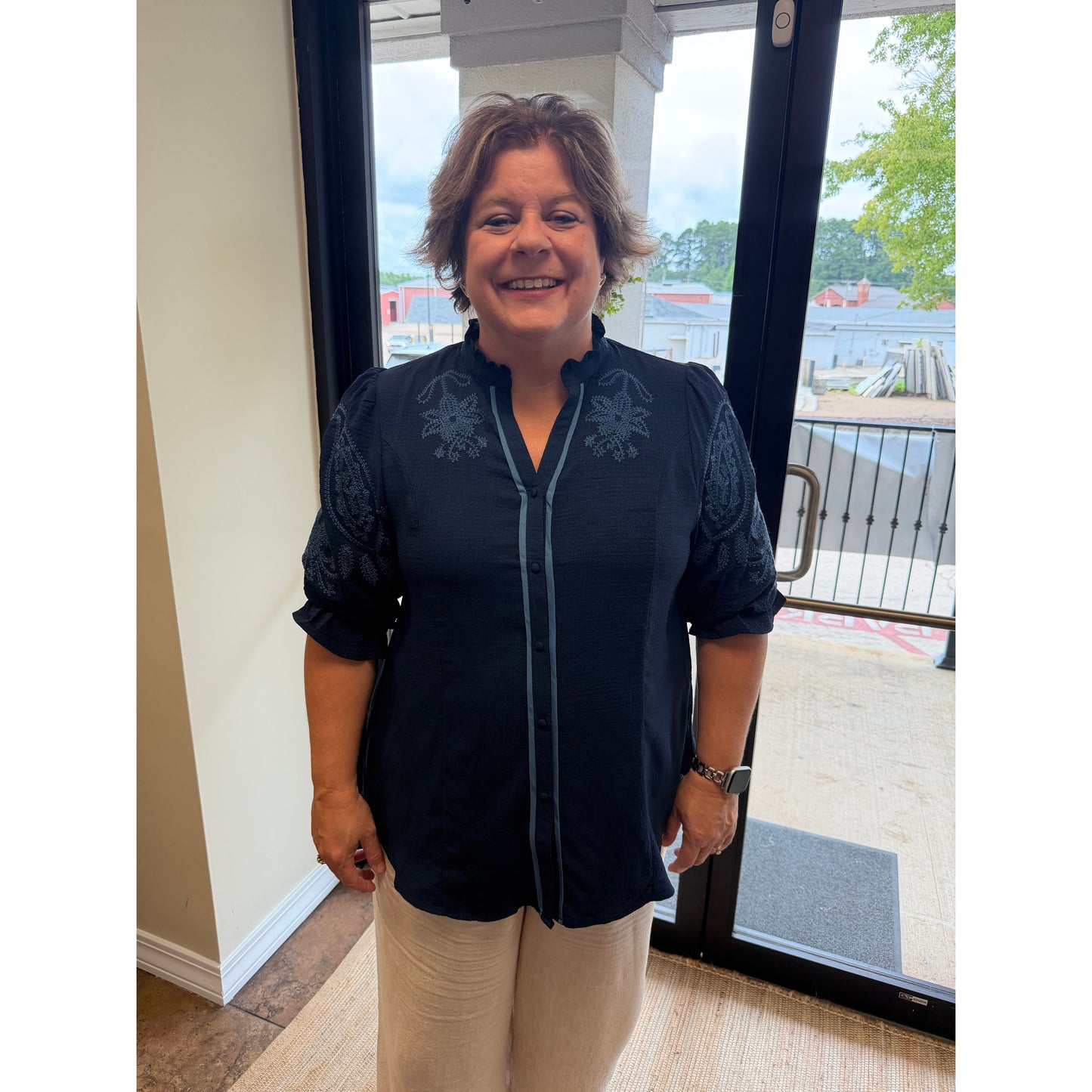 A woman smiling while wearing a dark navy blue embroidered blouse with beige pants, standing in front of a glass door.