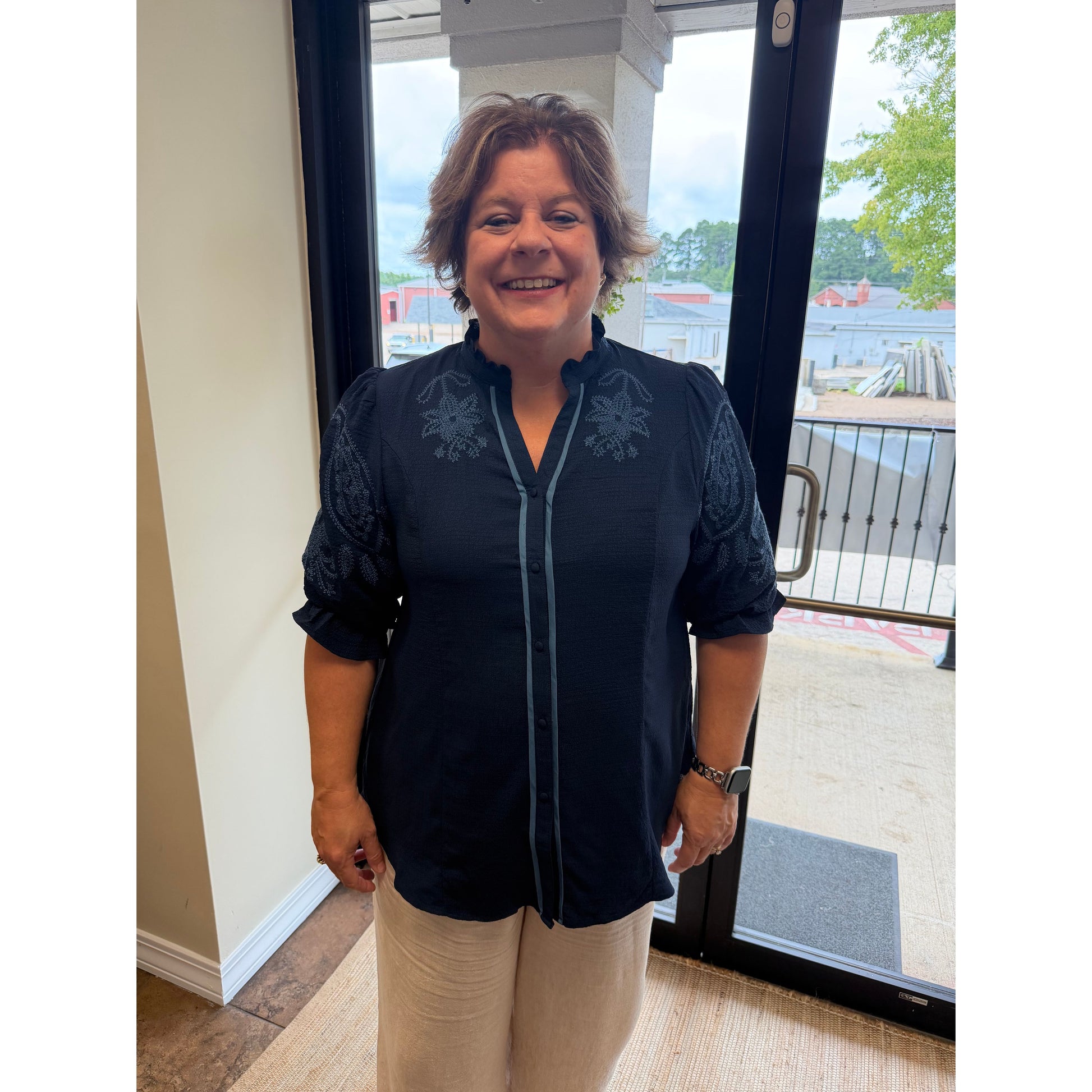 A woman smiling while wearing a dark navy blue embroidered blouse with beige pants, standing in front of a glass door.