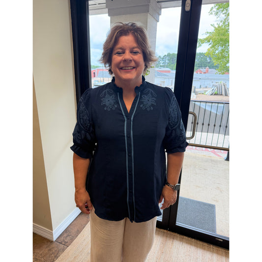 A woman smiling while wearing a dark navy blue embroidered blouse with beige pants, standing in front of a glass door.