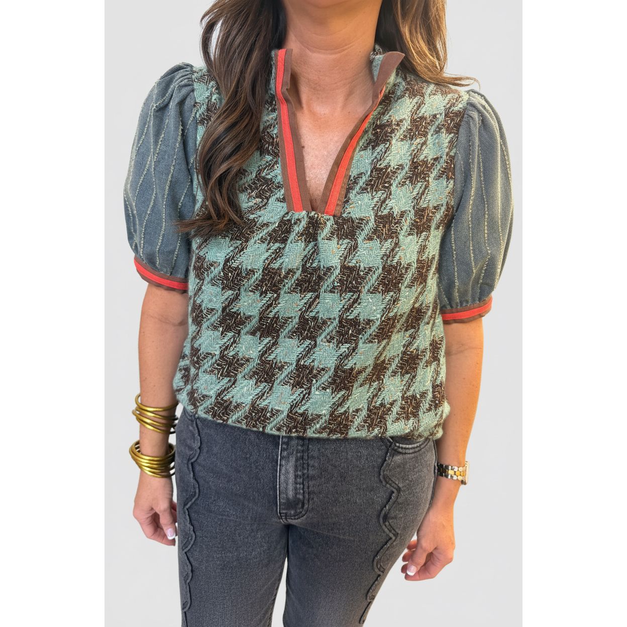 A woman wearing a short-sleeve houndstooth vest top with puff sleeves, featuring a brown and mint green pattern with orange-red trim on the collar and sleeve cuffs, paired with dark gray jeans.