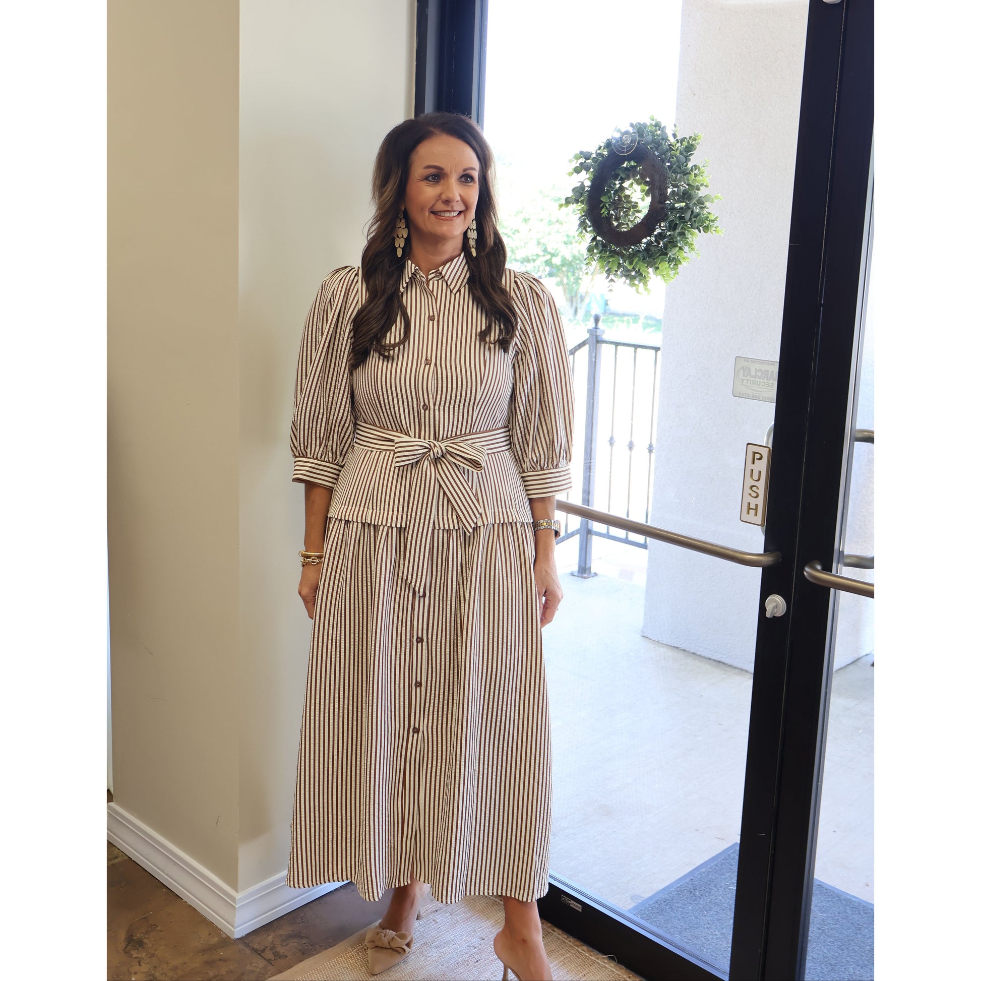 A woman wearing a beige and white vertically striped midi dress with puff sleeves, a button-down front, and a matching waist tie, standing in front of a glass door.