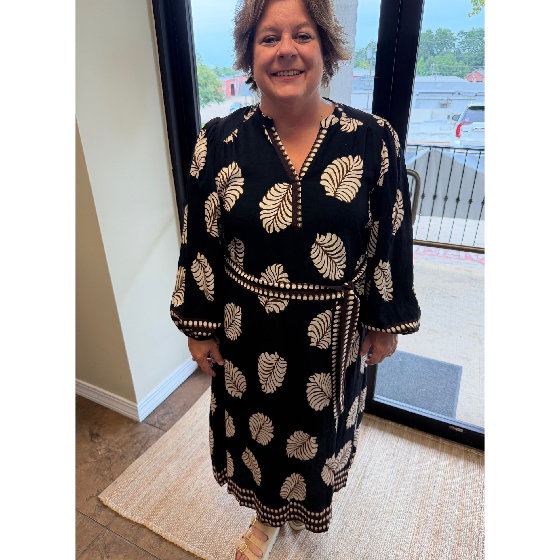 A woman wearing a black long-sleeve wrap dress with a beige palm leaf pattern, standing in front of a glass door.