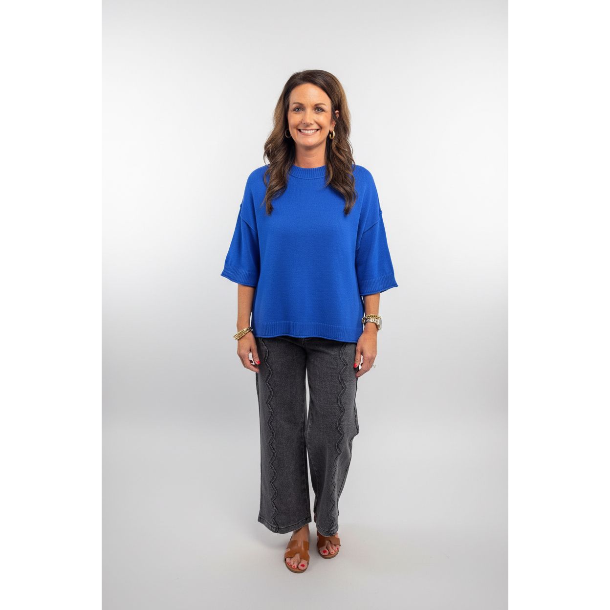 A woman modeling a bright blue short-sleeve knit sweater and dark gray textured wide-leg pants, standing indoors between two potted green topiary trees against a plain beige wall.