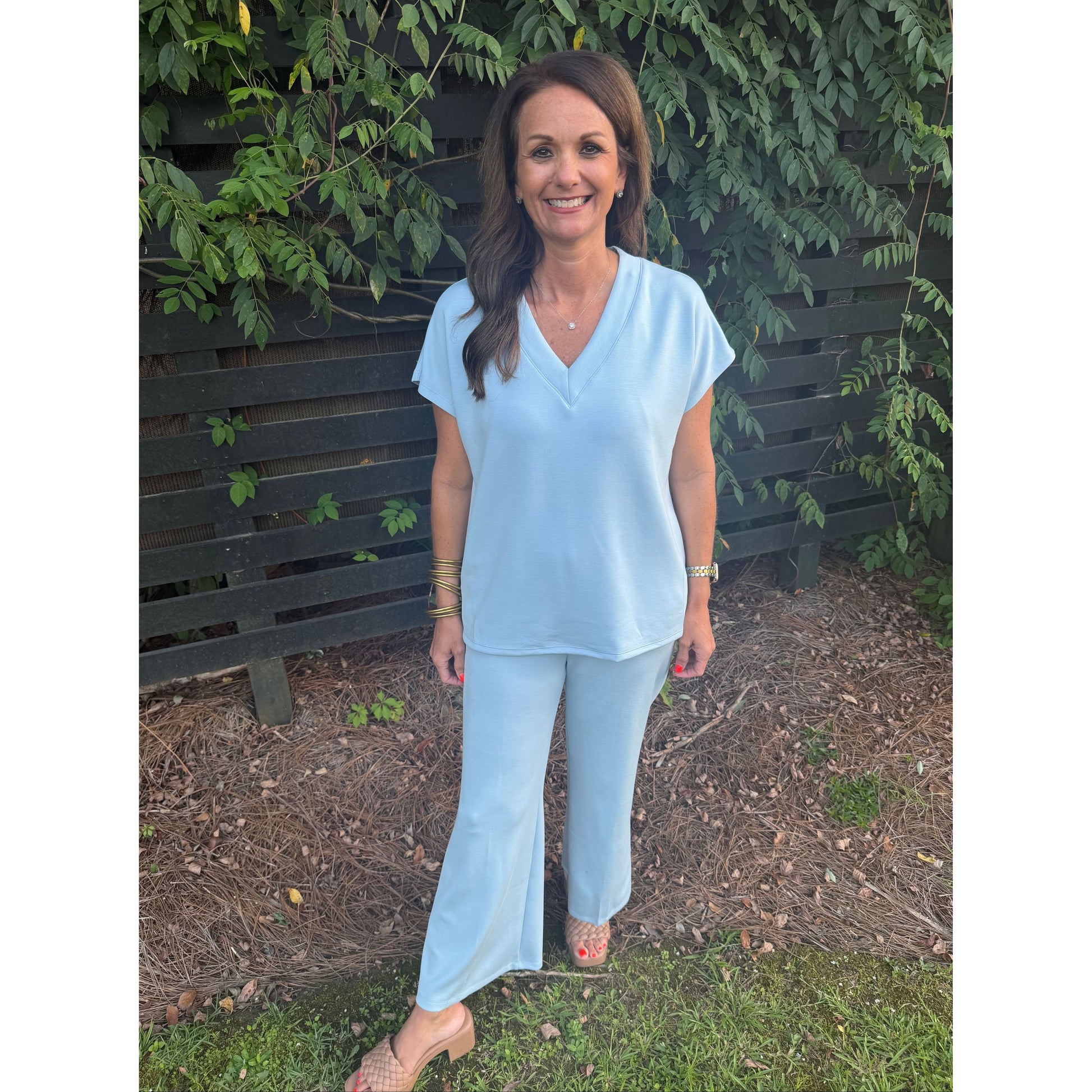 A woman wearing a light blue short-sleeve V-neck top and matching light blue wide-leg pants, standing outdoors in front of a dark wooden fence and green foliage.