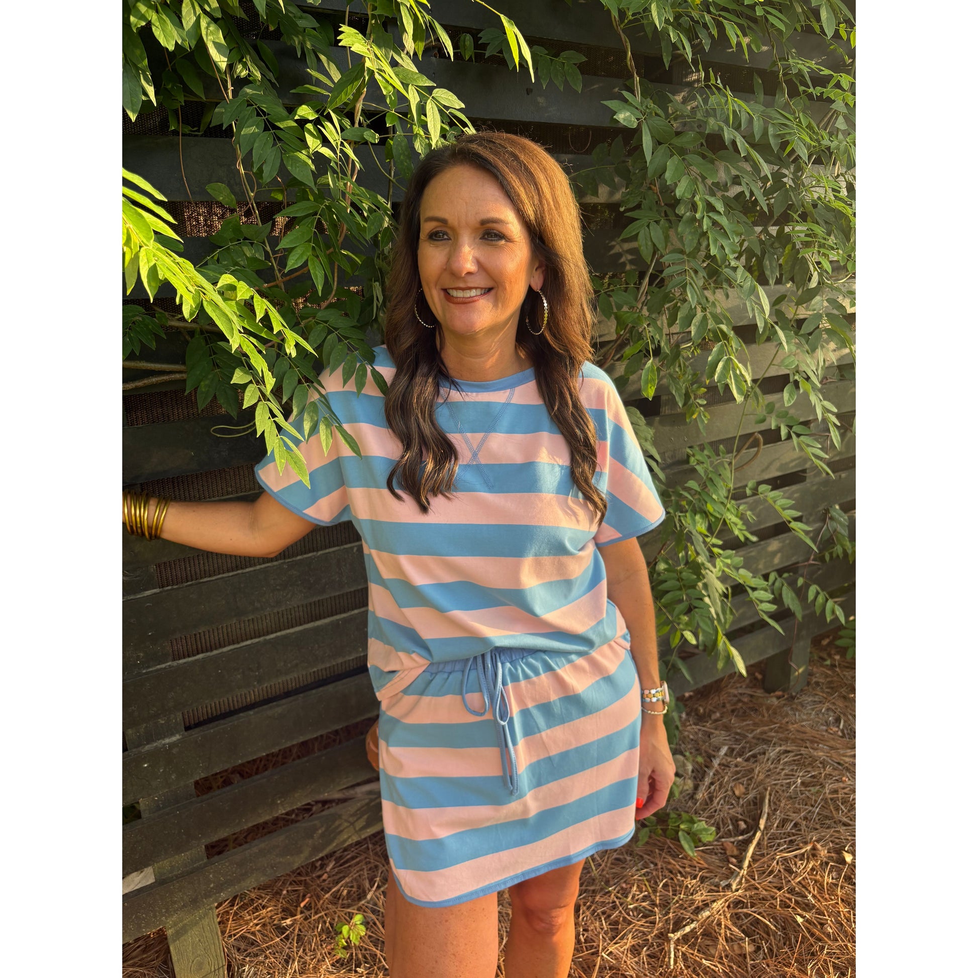 Woman wearing a pink and blue striped dress standing against a wooden wall with greenery.
