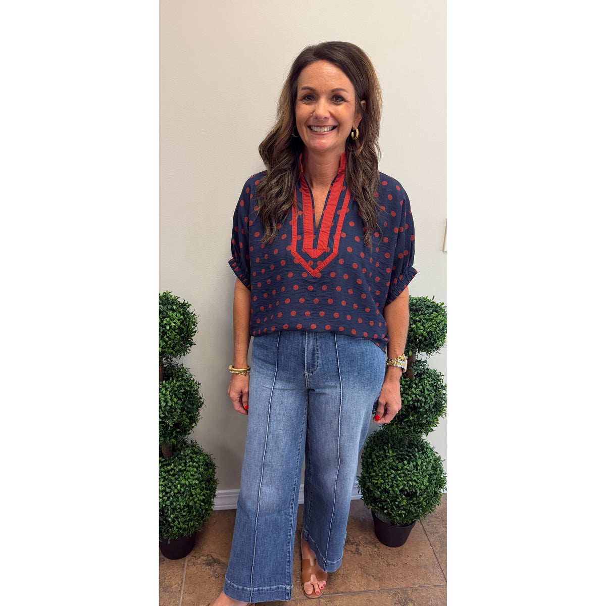 A woman modeling a navy blue short-sleeve top with red polka dots and a red V-neckline detail, paired with light blue wide-leg jeans, standing indoors between two green topiary trees.