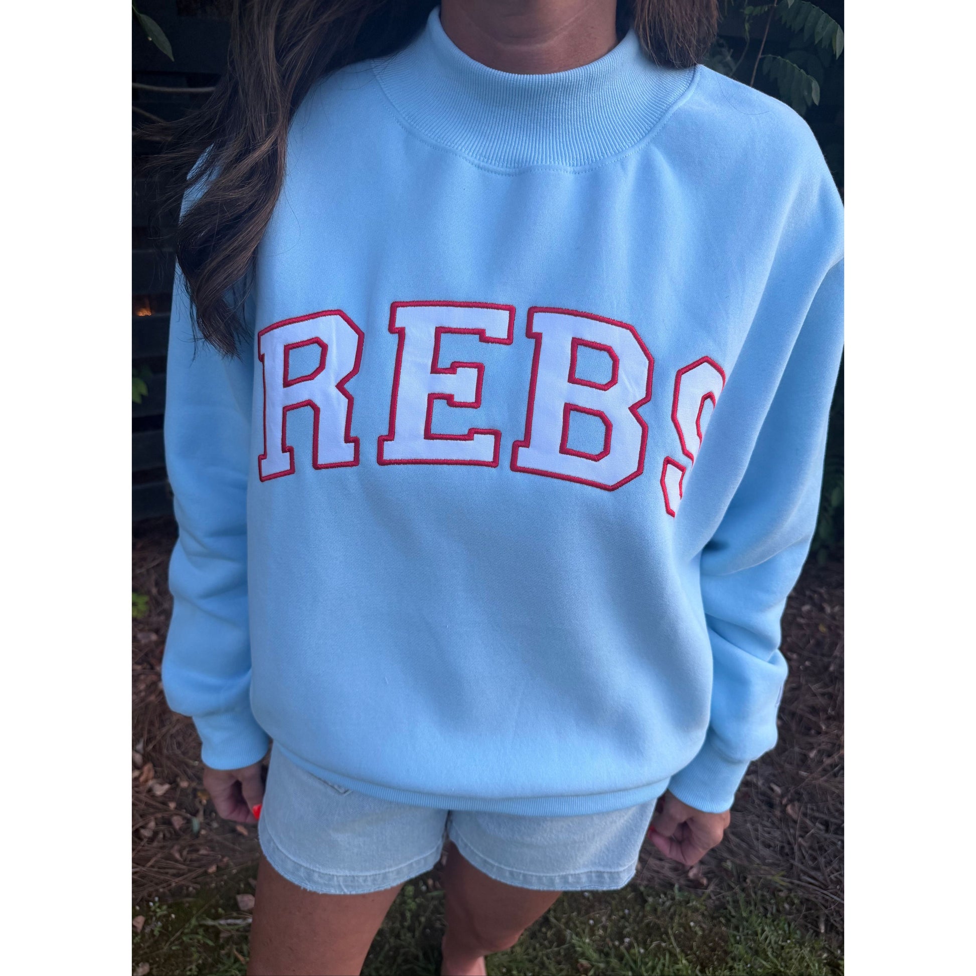A person wearing a light blue crewneck sweatshirt with 'REBS' embroidered in white with a red outline across the chest, paired with light denim shorts, standing outdoors.