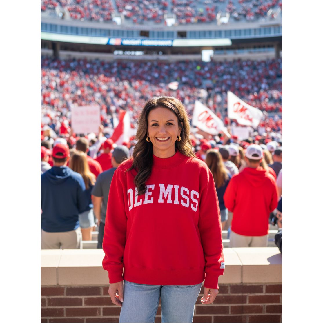 A woman smiling while wearing a bright red crewneck sweatshirt with 'OLE MISS' printed in white arched block letters across the chest, paired with light blue jeans, standing against a plain beige wall.