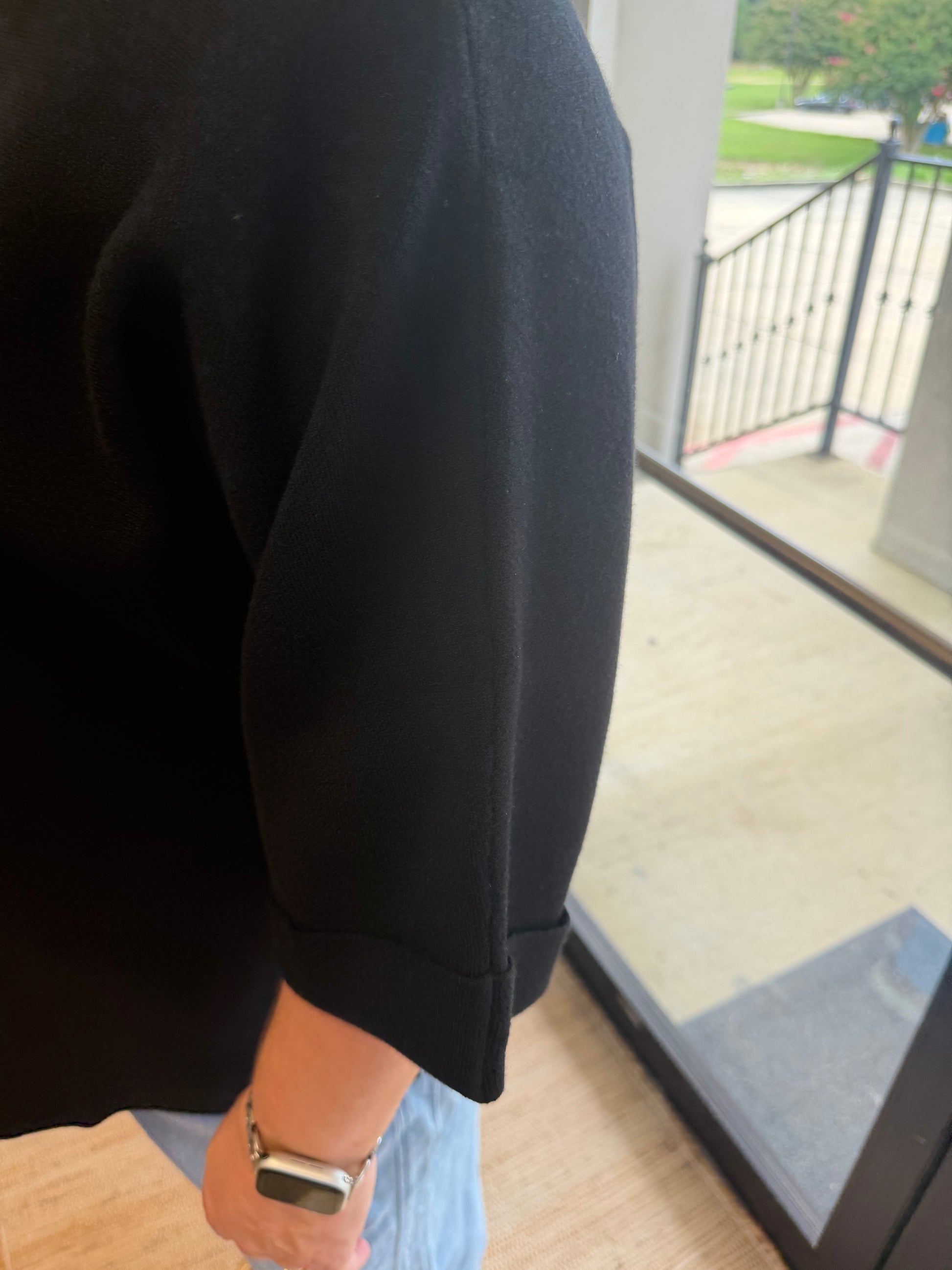A close-up of a person wearing a black short-sleeve top with a ribbed cuff, paired with light blue jeans and a silver smartwatch, standing indoors near a glass door.