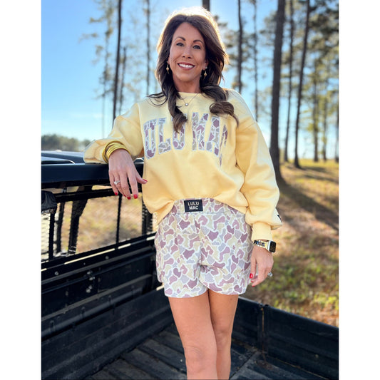 Woman wearing a yellow sweatshirt and patterned shorts standing in the back of a truck with trees in the background.
