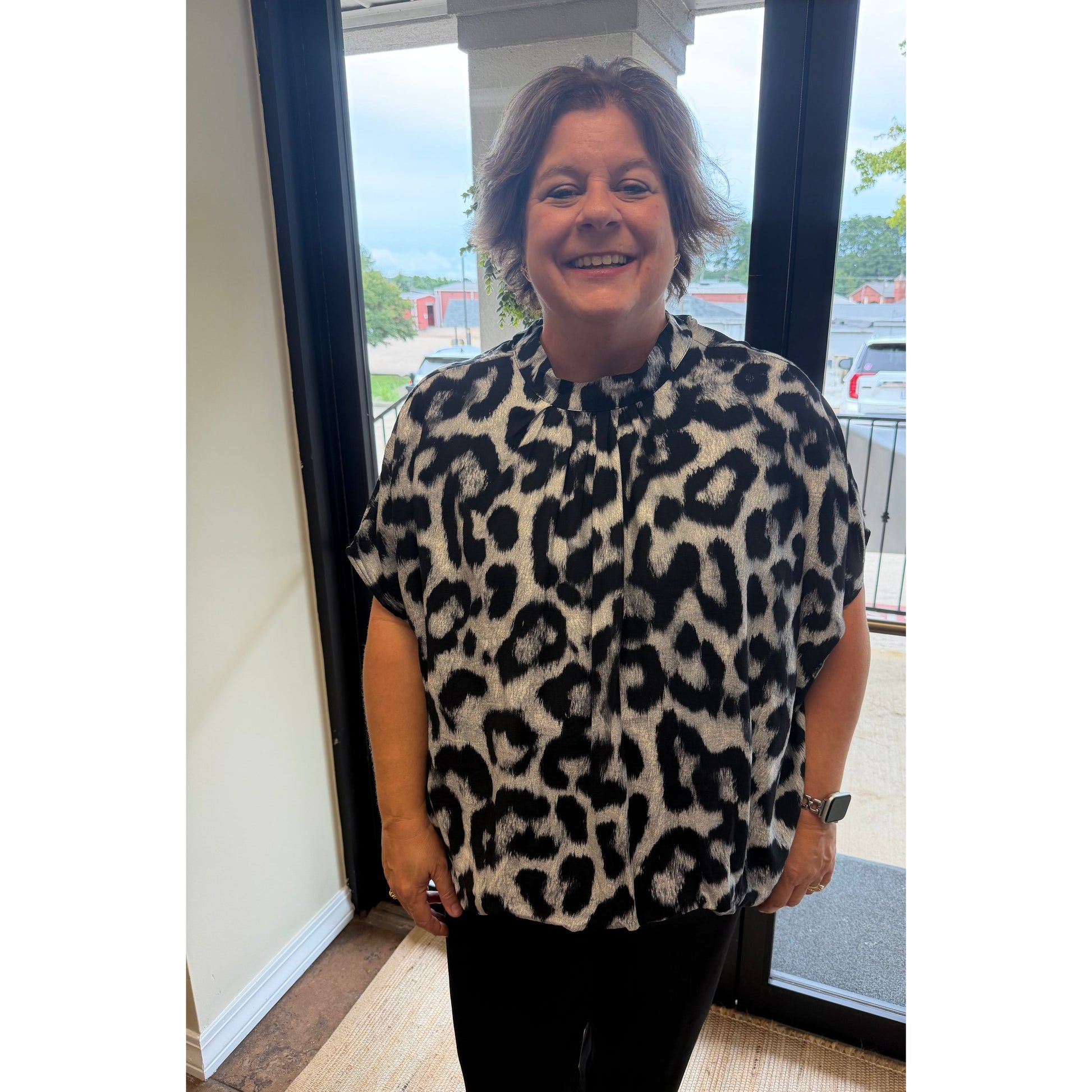 A woman smiling while wearing a short-sleeve leopard print blouse with black pants, standing in front of a glass door.
