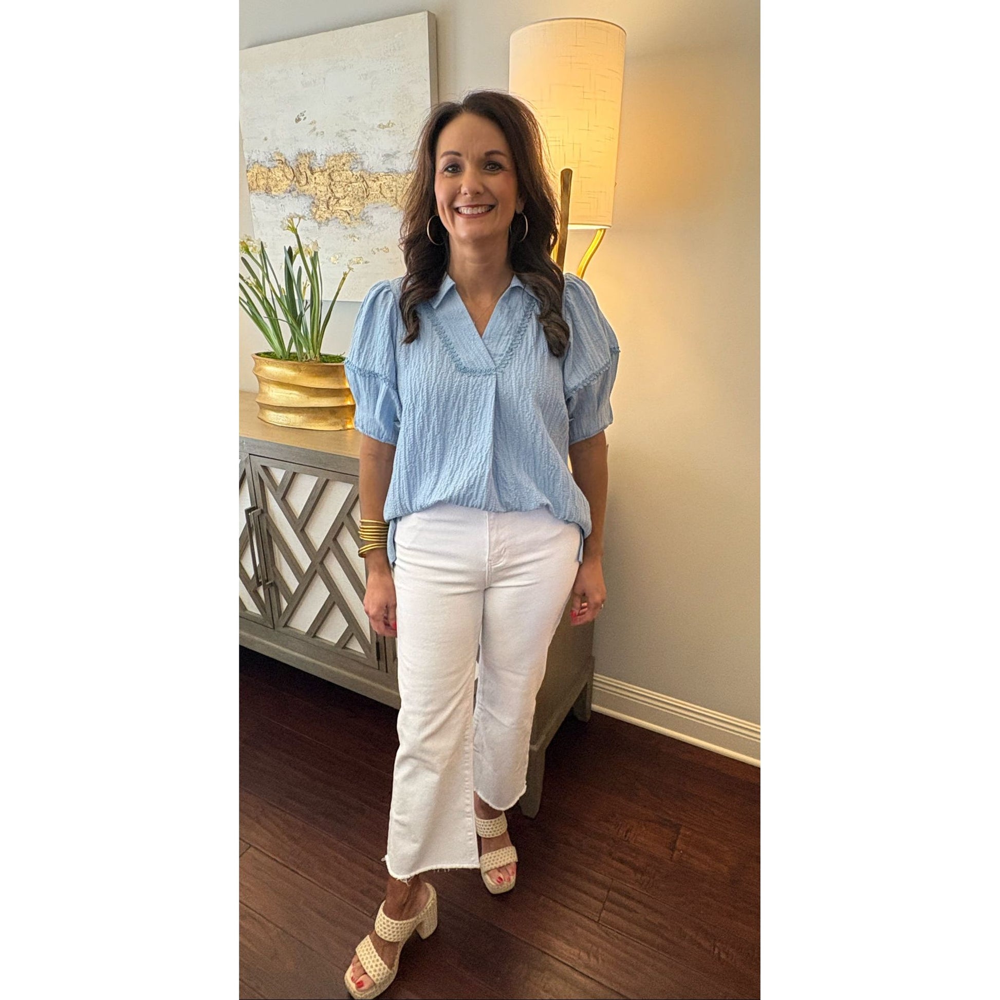 Woman in a blue top and white pants standing in a room with lamps and decor.