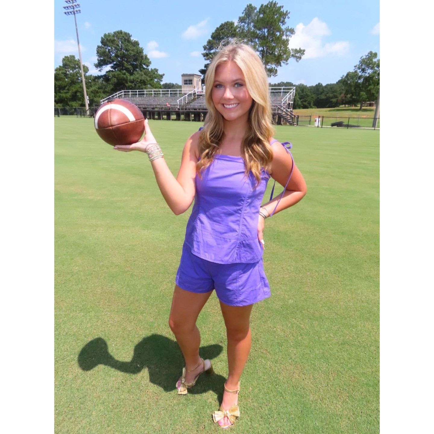 A woman stands on a grassy field holding a football, wearing a matching purple sleeveless top and shorts set with beige sandals.