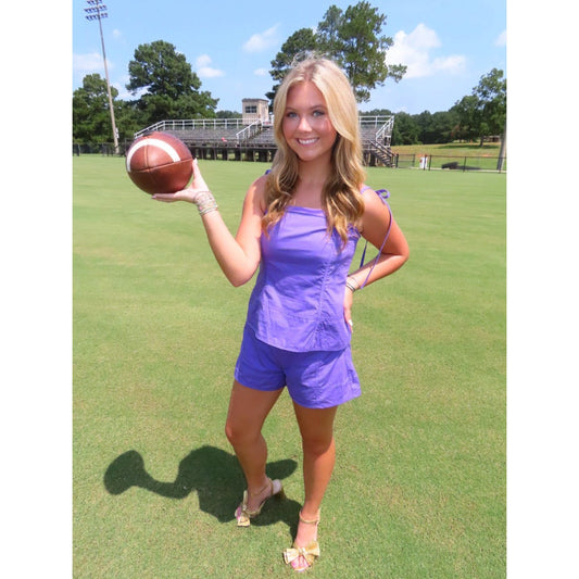 A woman stands on a grassy field holding a football, wearing a matching purple sleeveless top and shorts set with beige sandals.
