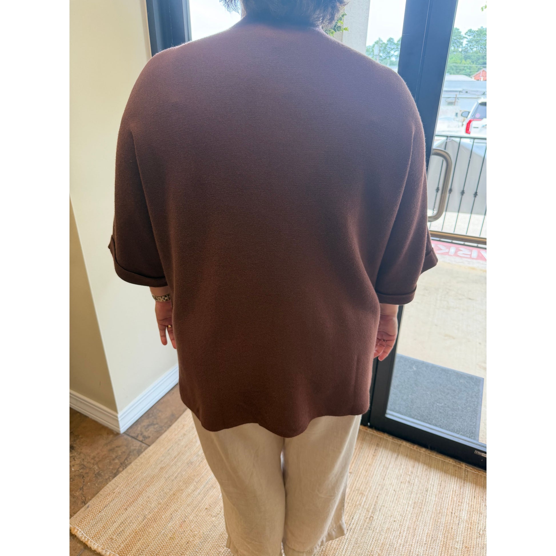 A person viewed from behind wearing a brown short-sleeve knit top and light beige pants, standing indoors near a glass door.