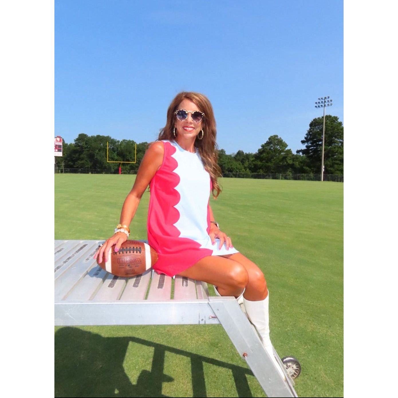 A woman wearing a sleeveless white and pink dress sits on a metal bleacher at a football field, holding a football under her arm. The background shows a green grass field, trees, and a blue sky.