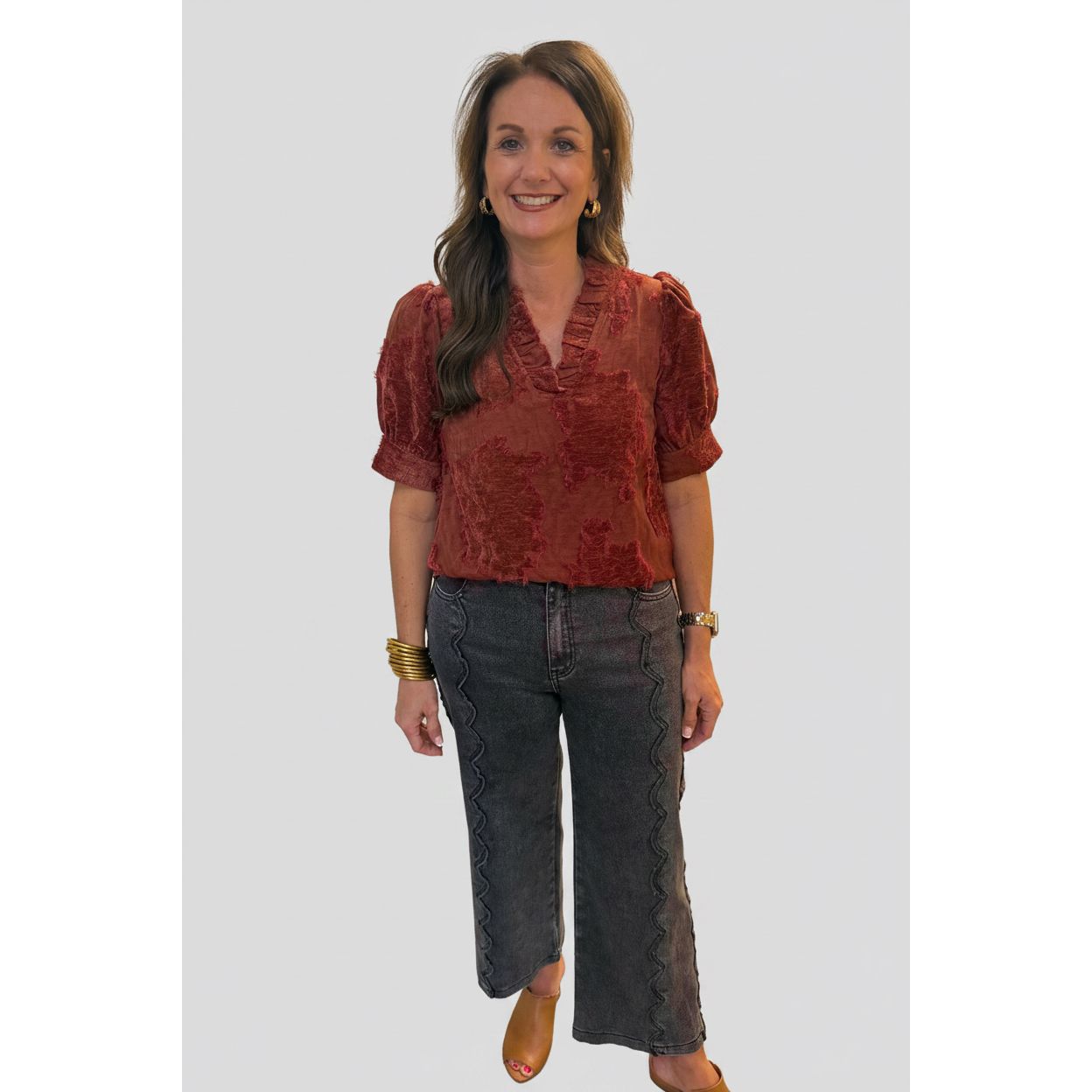 A woman posing indoors wearing a rust-red textured short-sleeve blouse and dark gray cropped pants with decorative stitching.