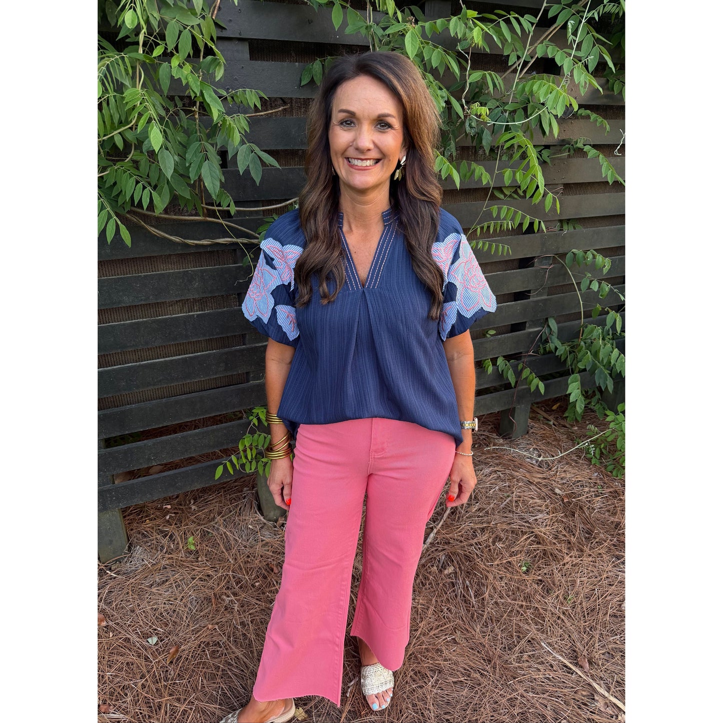 Woman wearing a blue top with floral sleeves and pink pants standing outdoors.