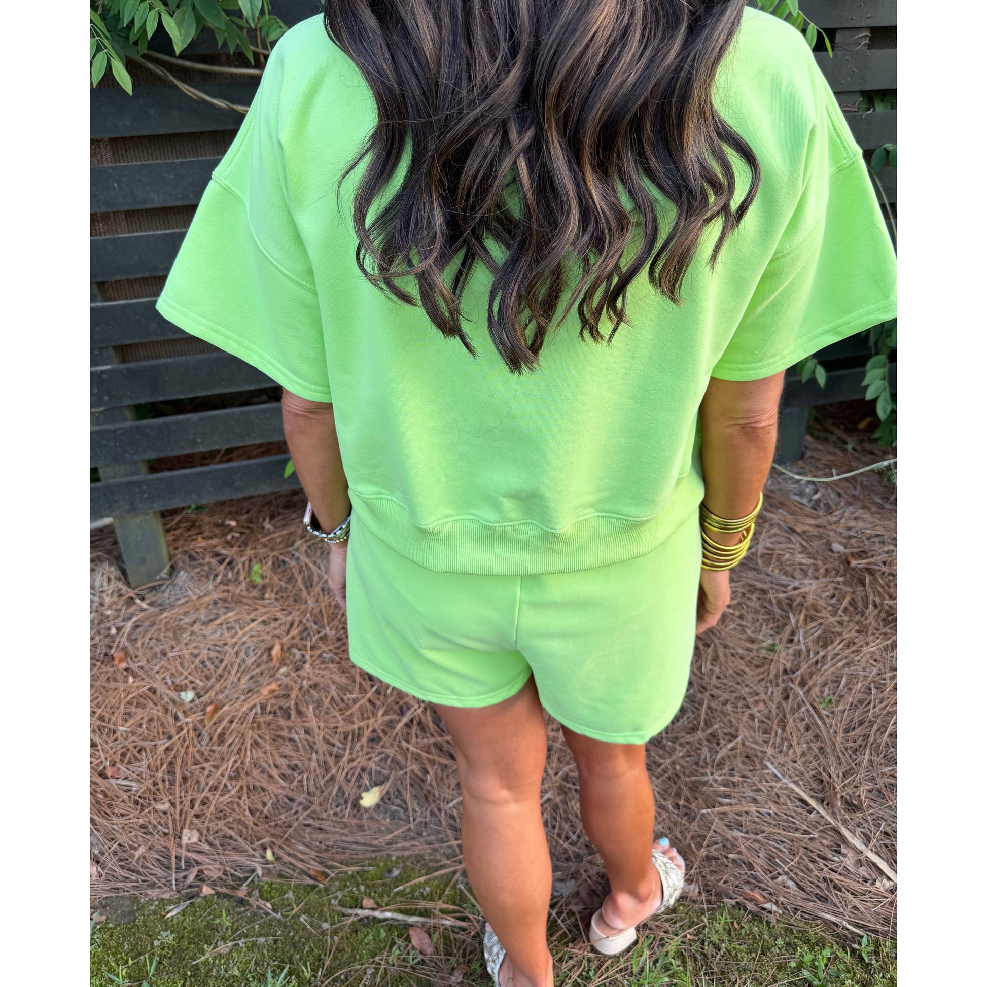 Person wearing a bright green outfit standing outdoors with a wooden fence and plants in the background.