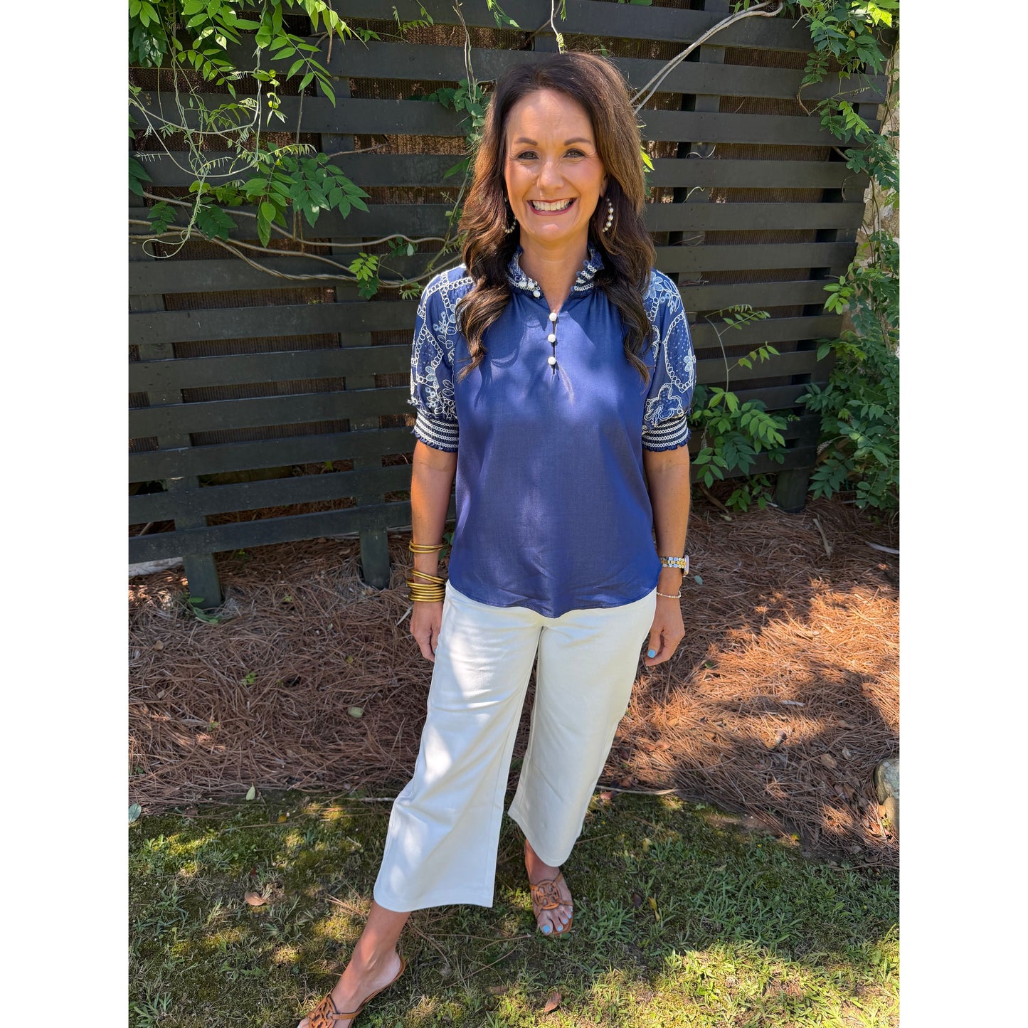 Woman wearing a blue patterned shirt and white pants standing in front of a wooden fence with greenery.