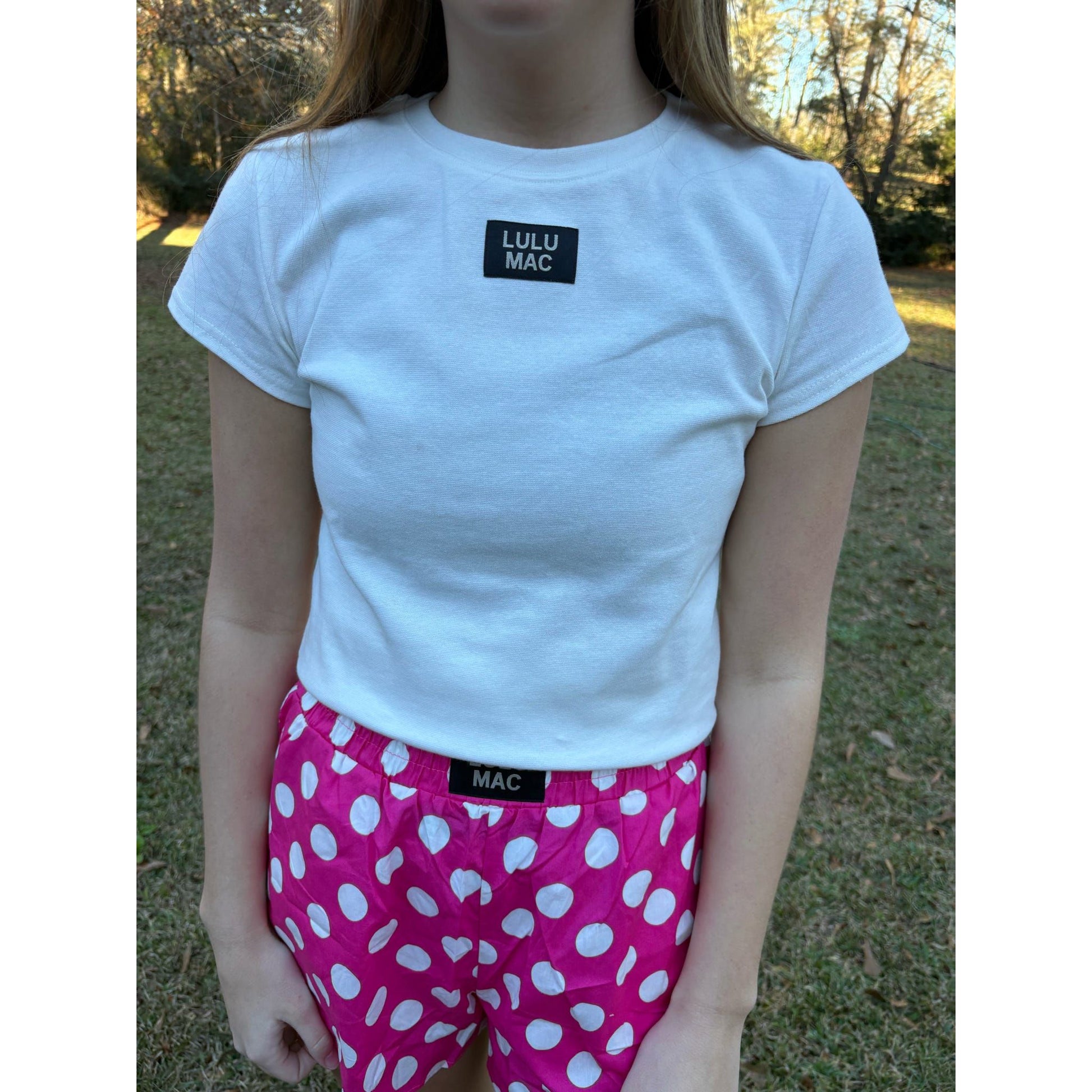 Person wearing a white t-shirt with 'Lulu Mac' logo and pink polka dot shorts outdoors.