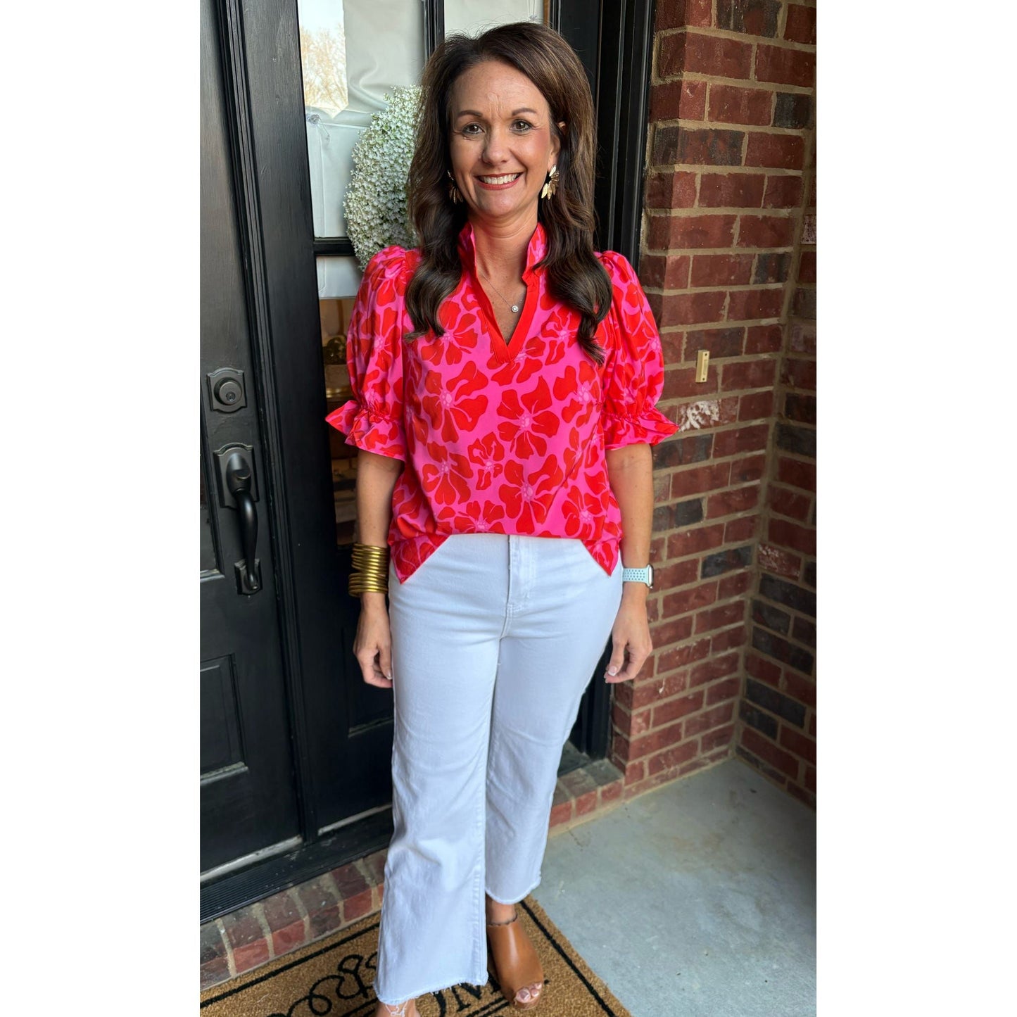 Woman wearing a red floral blouse and white pants standing in front of a brick wall.