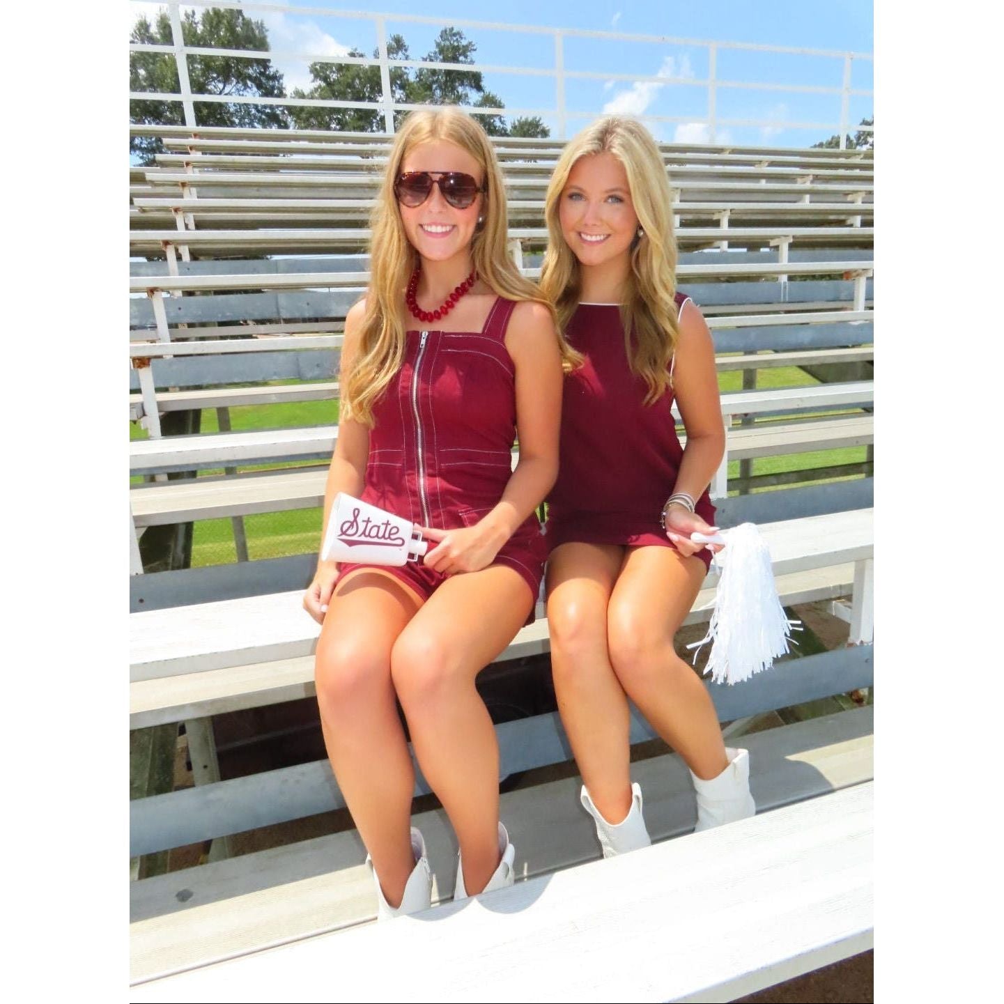 Two women sitting together on outdoor stadium bleachers wearing matching sleeveless burgundy mini dresses and white cowboy boots, with one holding a white 'State' sign and the other holding a white tassel.