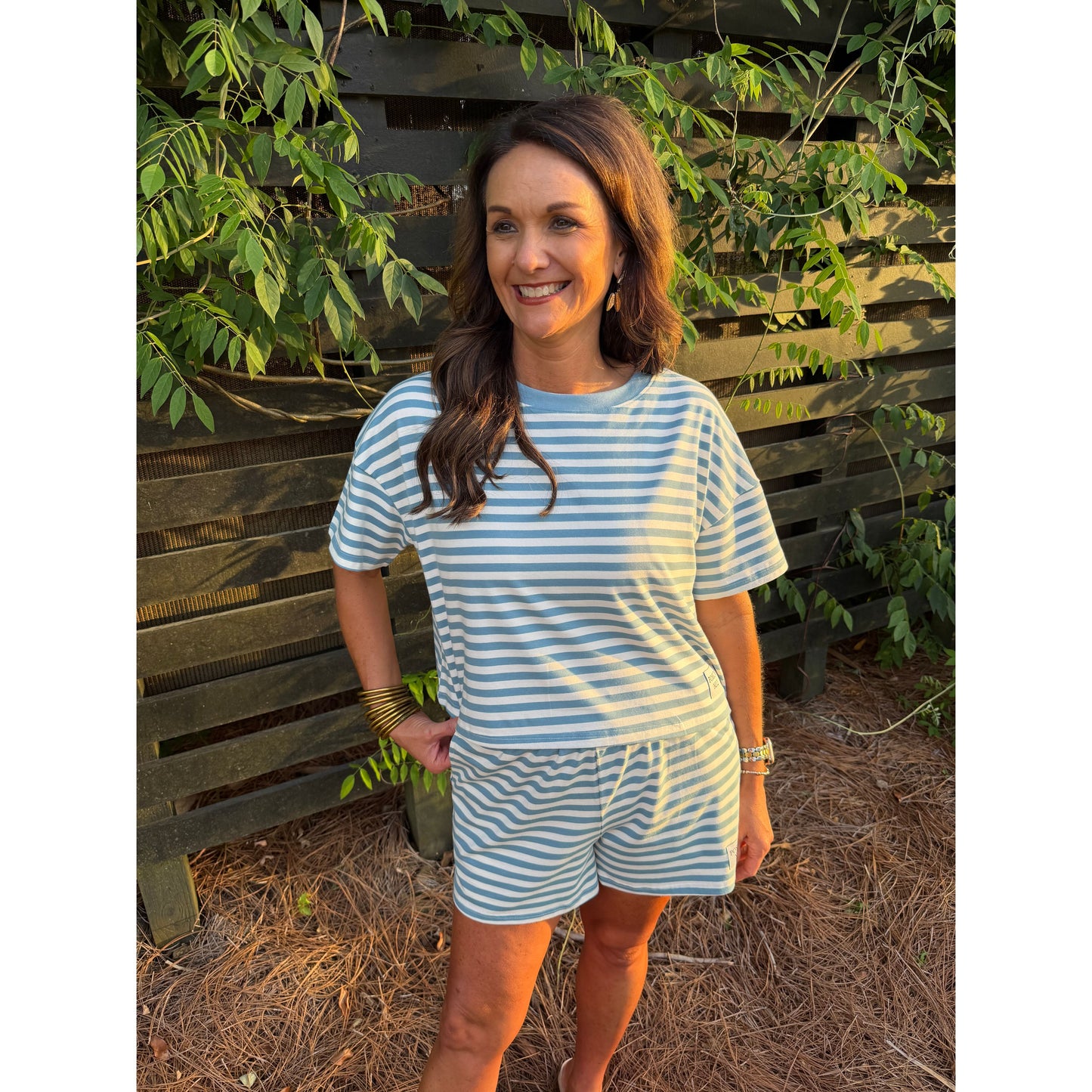 Woman wearing a blue and white striped outfit standing in front of a wooden fence with greenery.