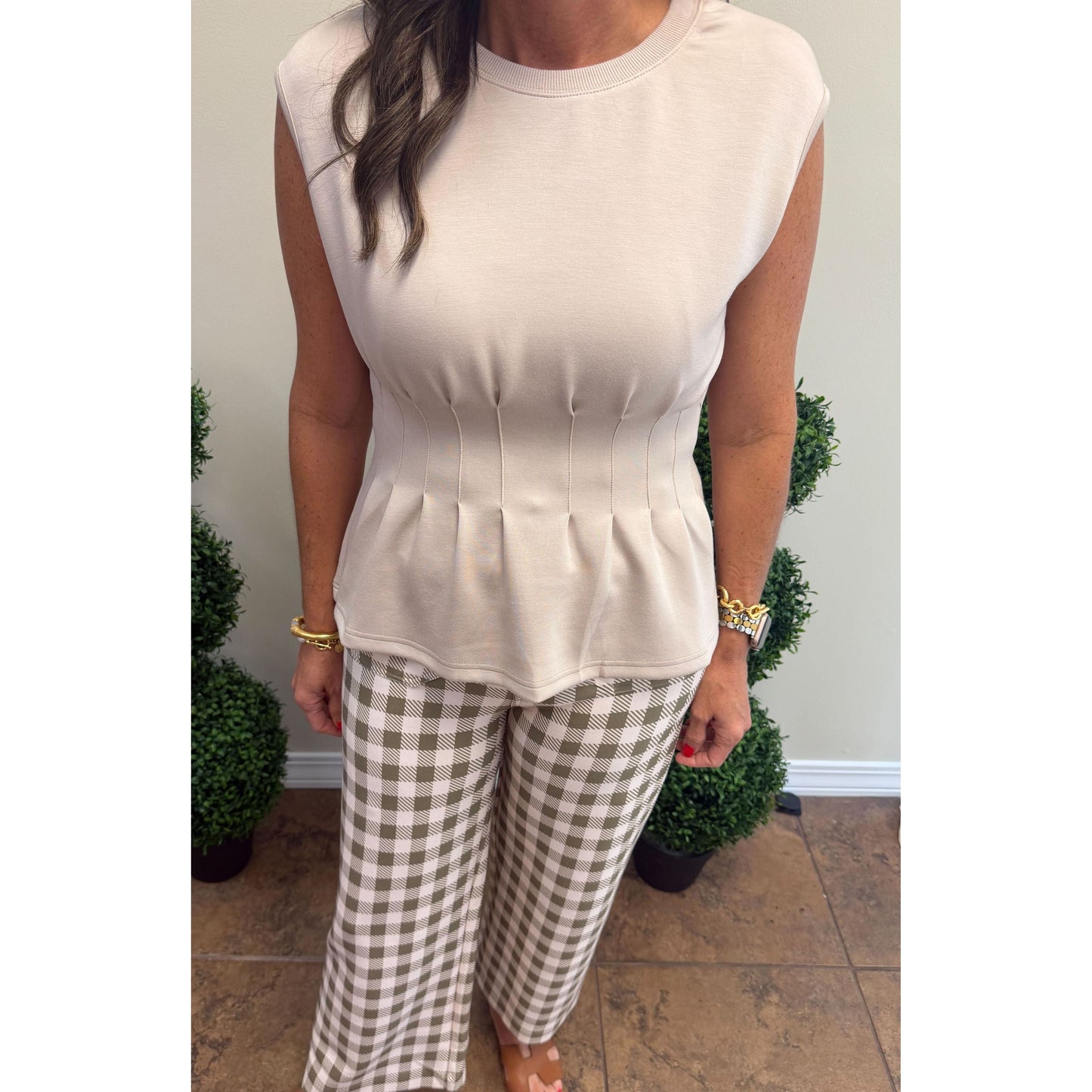 A woman modeling a sleeveless beige top with a structured corset-style waist and pleated peplum hem, paired with brown and beige gingham wide-leg pants, standing indoors.